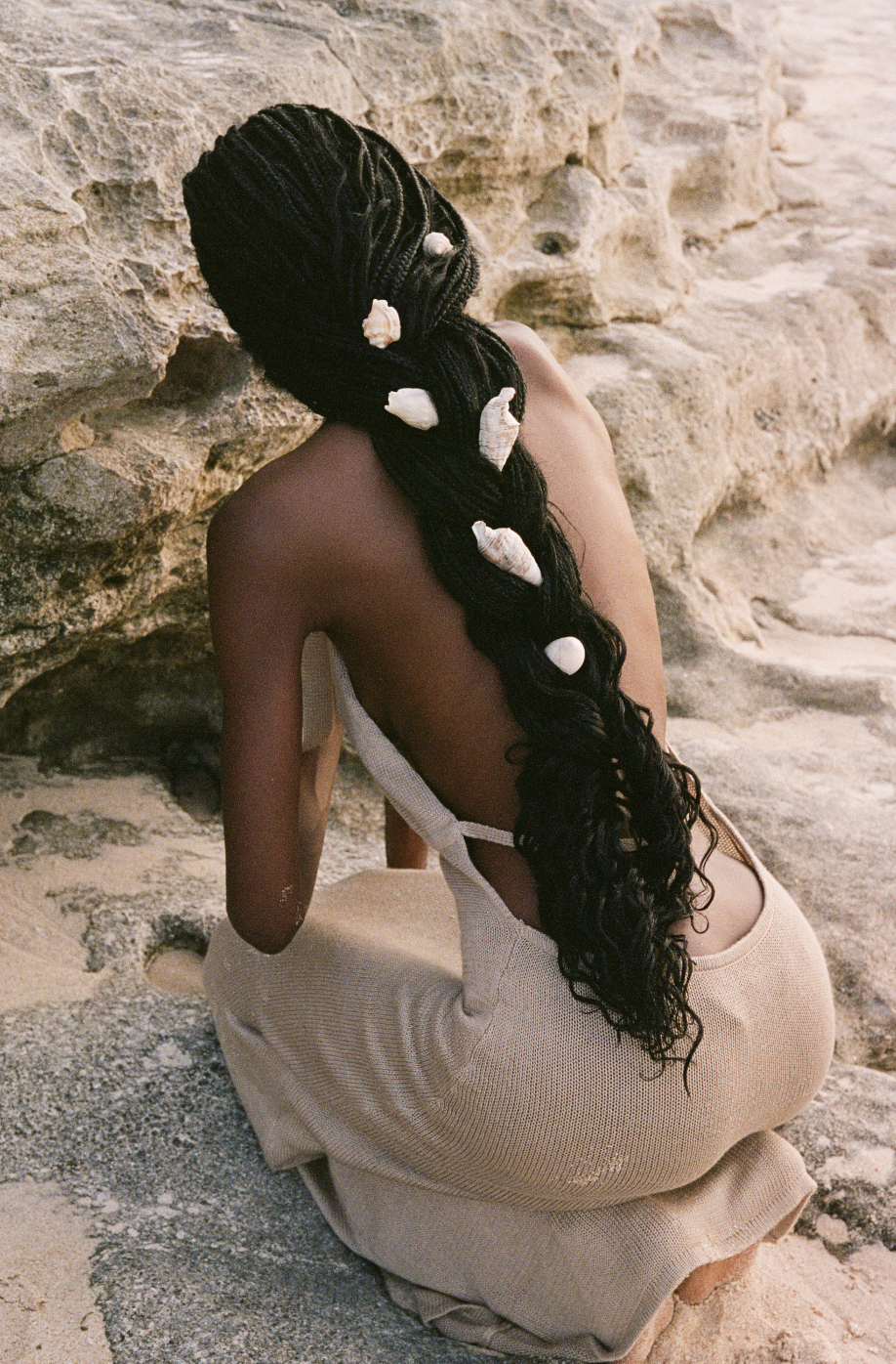 A person with long braided hair decorated with shells, wearing a beige dress, sitting on rock formations at the beach.