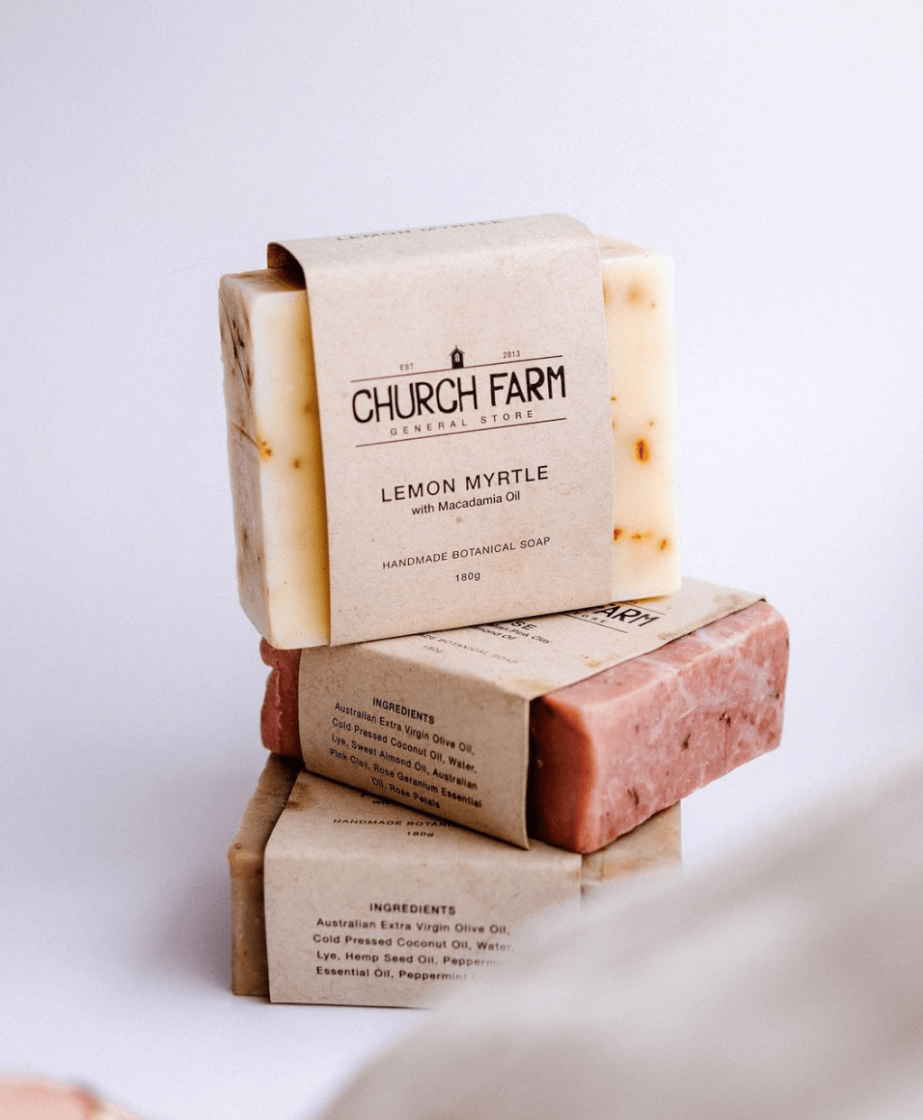 Stack of three handmade botanical soap bars with labels, including a top bar labeled as "Church Farm General Store Lemon Myrtle with Macadamia Oil."