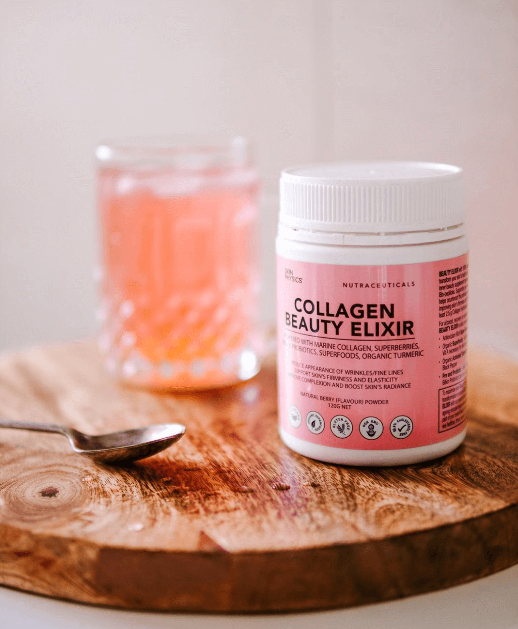 A jar of Collagen Beauty Elixir on a wooden board with a spoon and a glass of pink liquid in the background.
