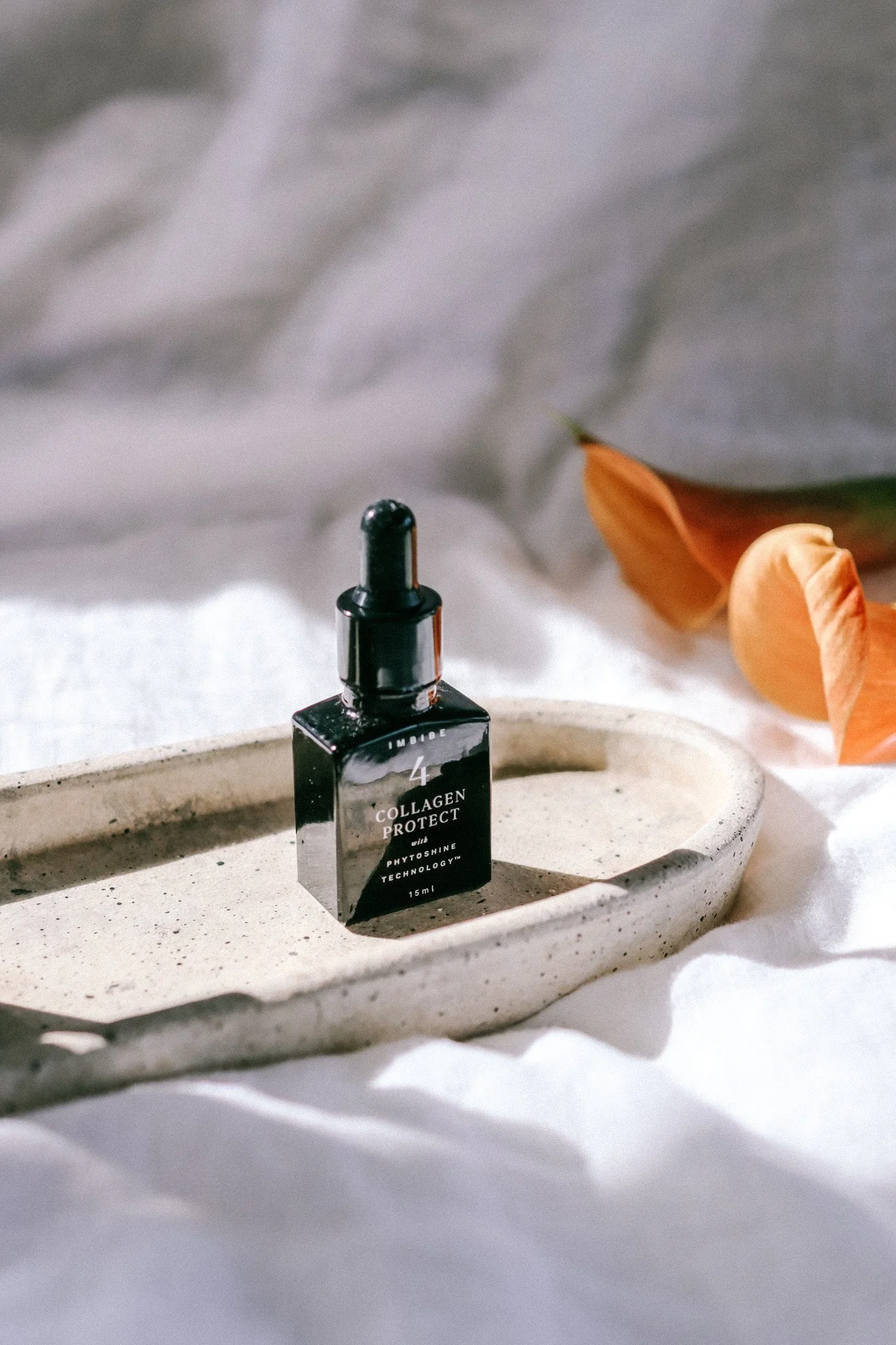 A small black dropper bottle labeled "Collagen Protect" with Phytoshine Technology, placed on a ceramic tray with an orange flower nearby, on a white fabric background.