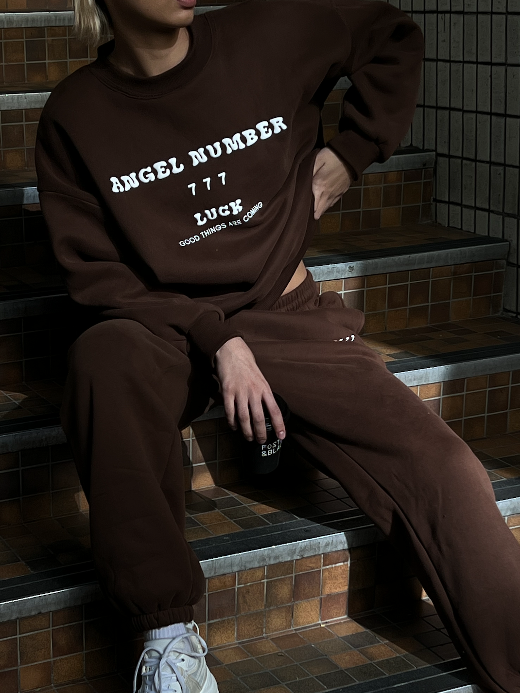 Person sitting on stairs wearing a brown tracksuit with 'Angel Number 777 Luck Good Things are Coming' text on the sweatshirt, holding a small black object.