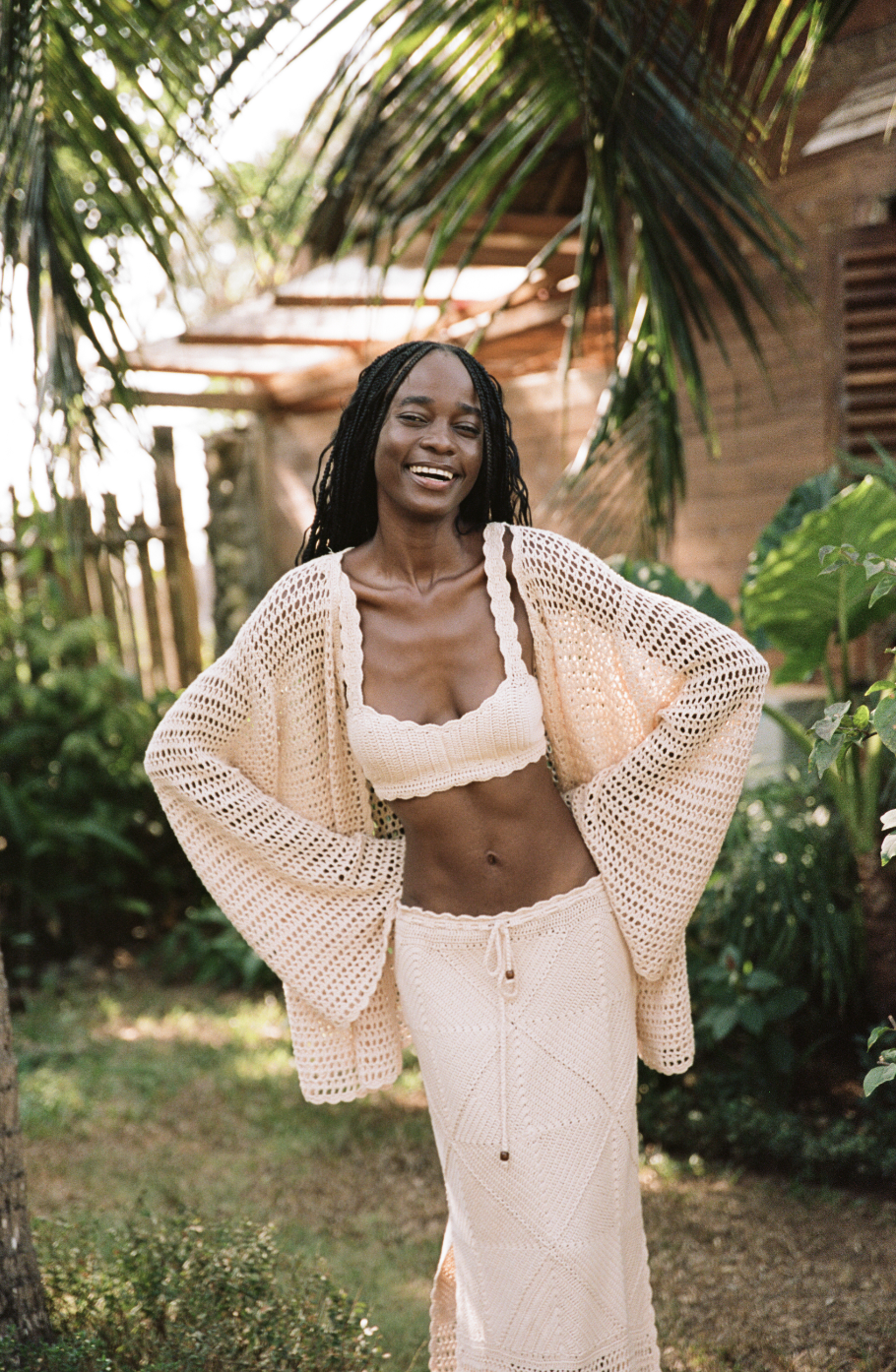 Person smiling in a crochet outfit standing outdoors near tropical plants and a wooden structure.
