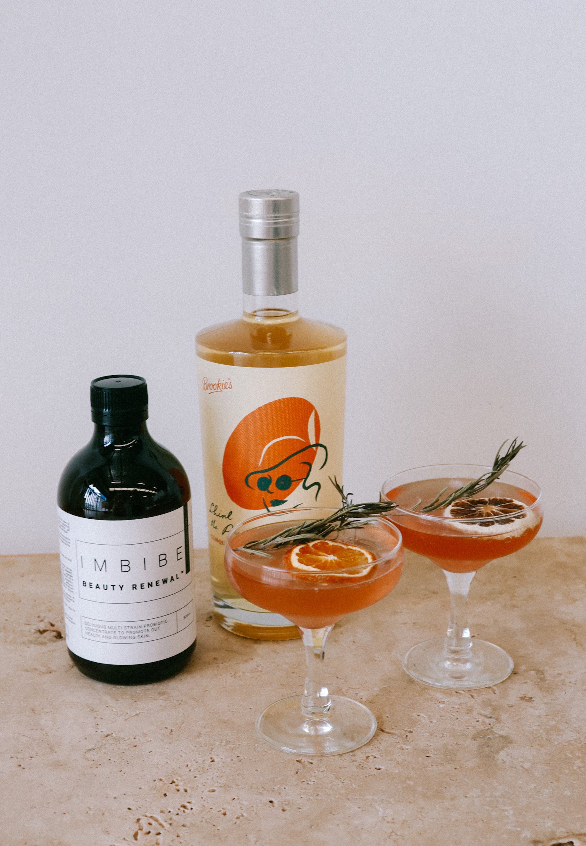 Two cocktail glasses with garnished drinks alongside a bottle of Brookie’s gin and a bottle of Imbibe Beauty Renewal drink, on a marble surface.