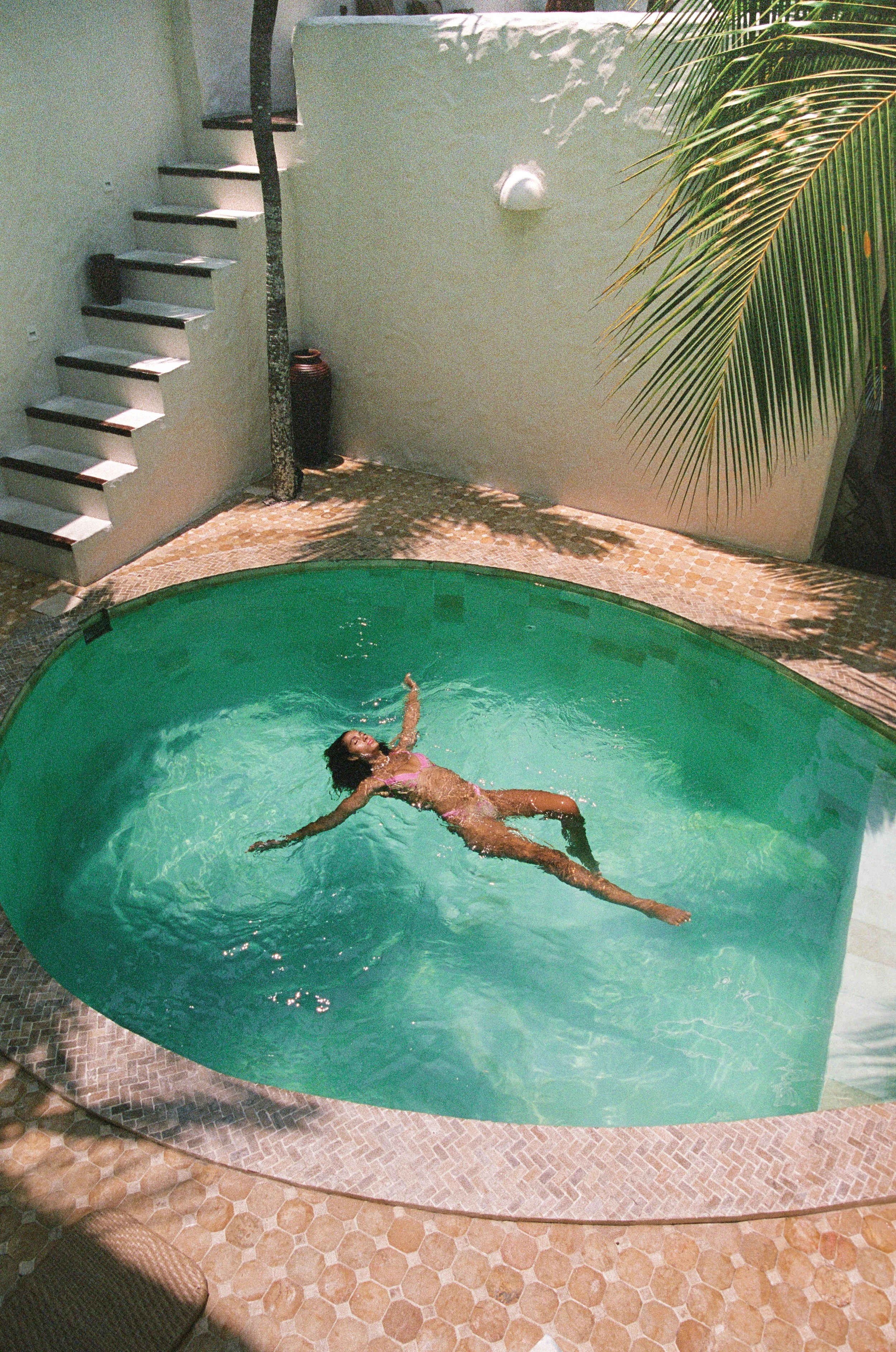 Person floating in a small backyard pool surrounded by a tiled patio and palm leaves.