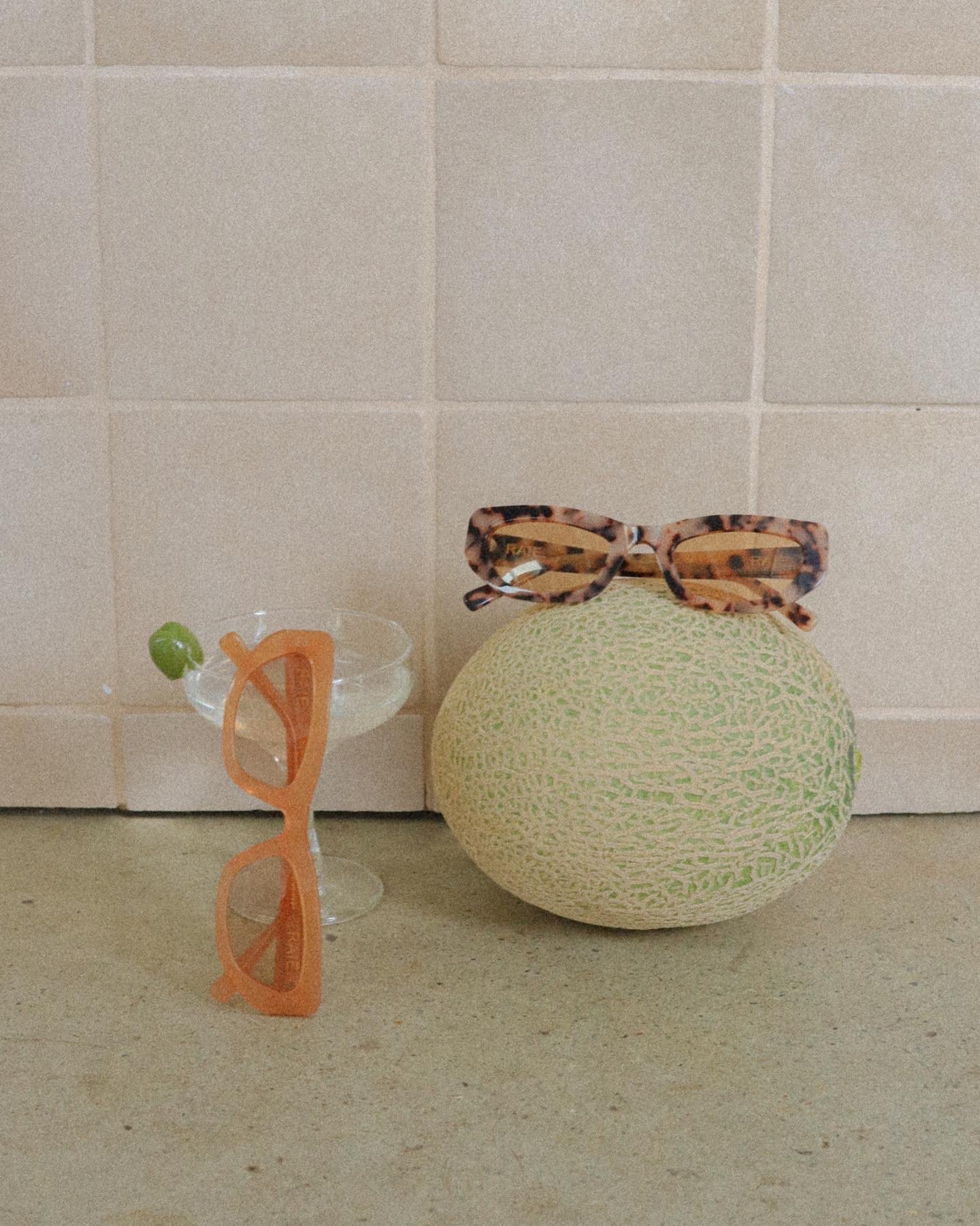 A cantaloupe with sunglasses resting on top, next to a glass filled with liquid and an olive, and a pair of orange sunglasses leaning against the glass, all set against a tiled wall.