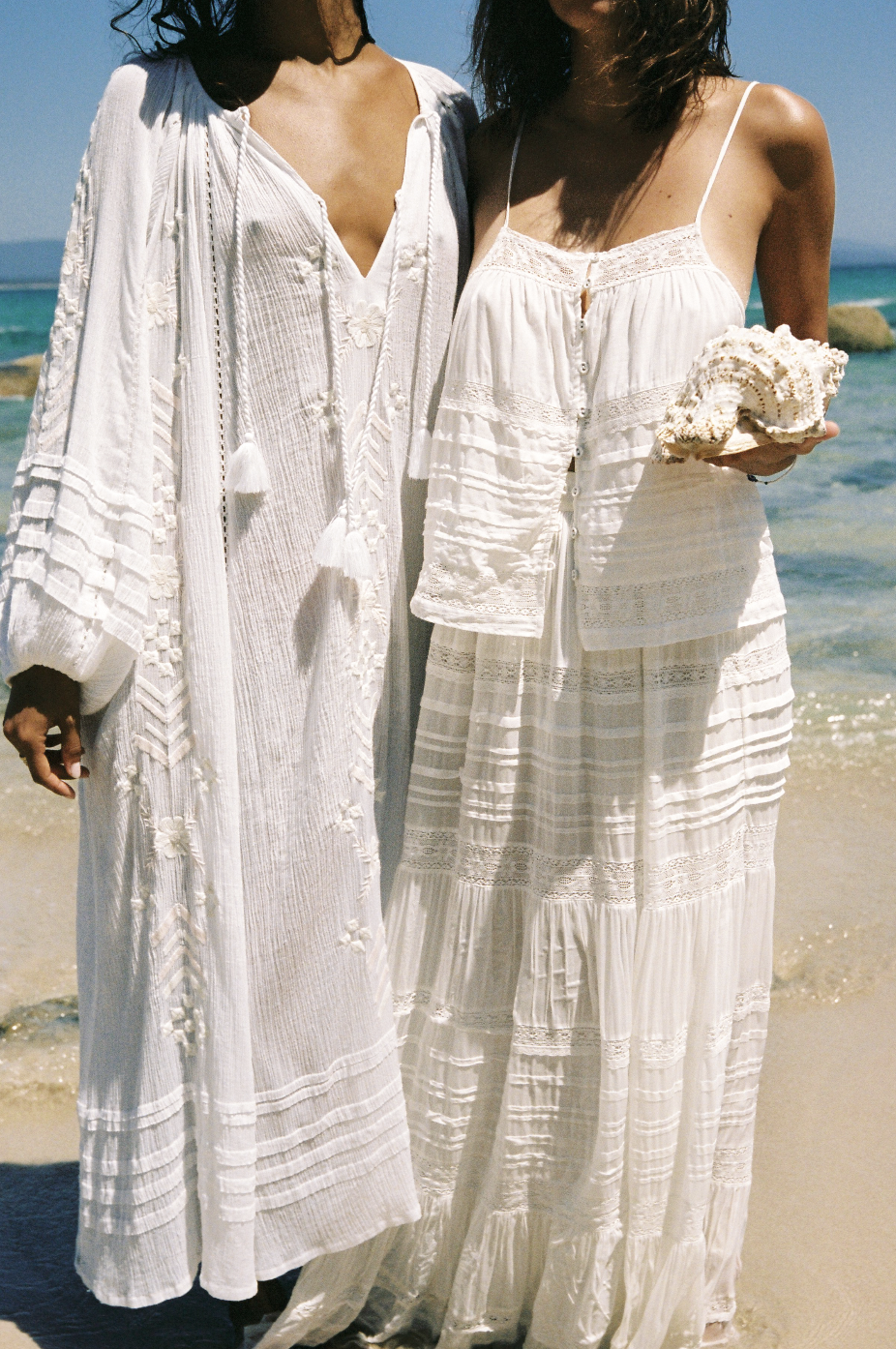Two women wearing white bohemian-style dresses on a beach, one holding a seashell. The background shows the ocean and blue sky.