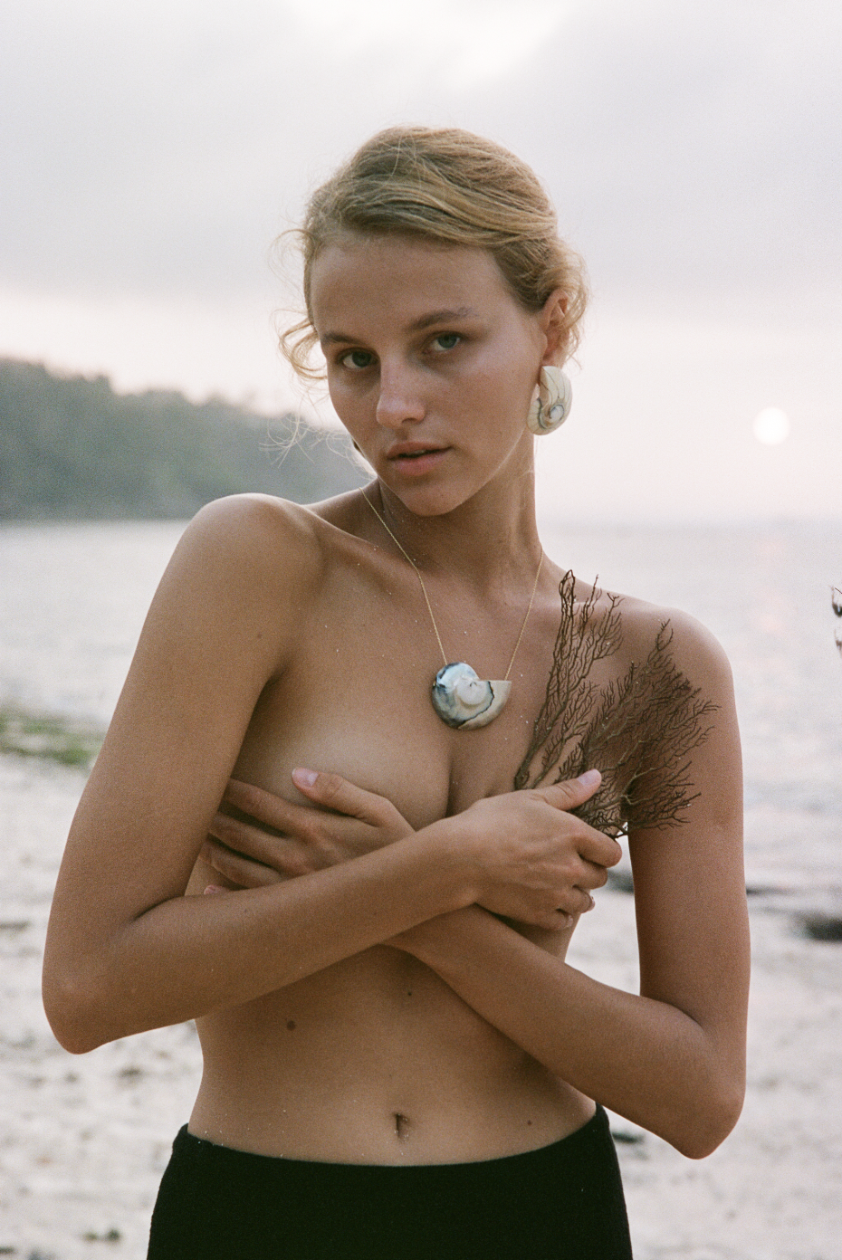 Person standing on a beach, wearing a shell necklace and earrings, holding a piece of coral, with the ocean in the background.