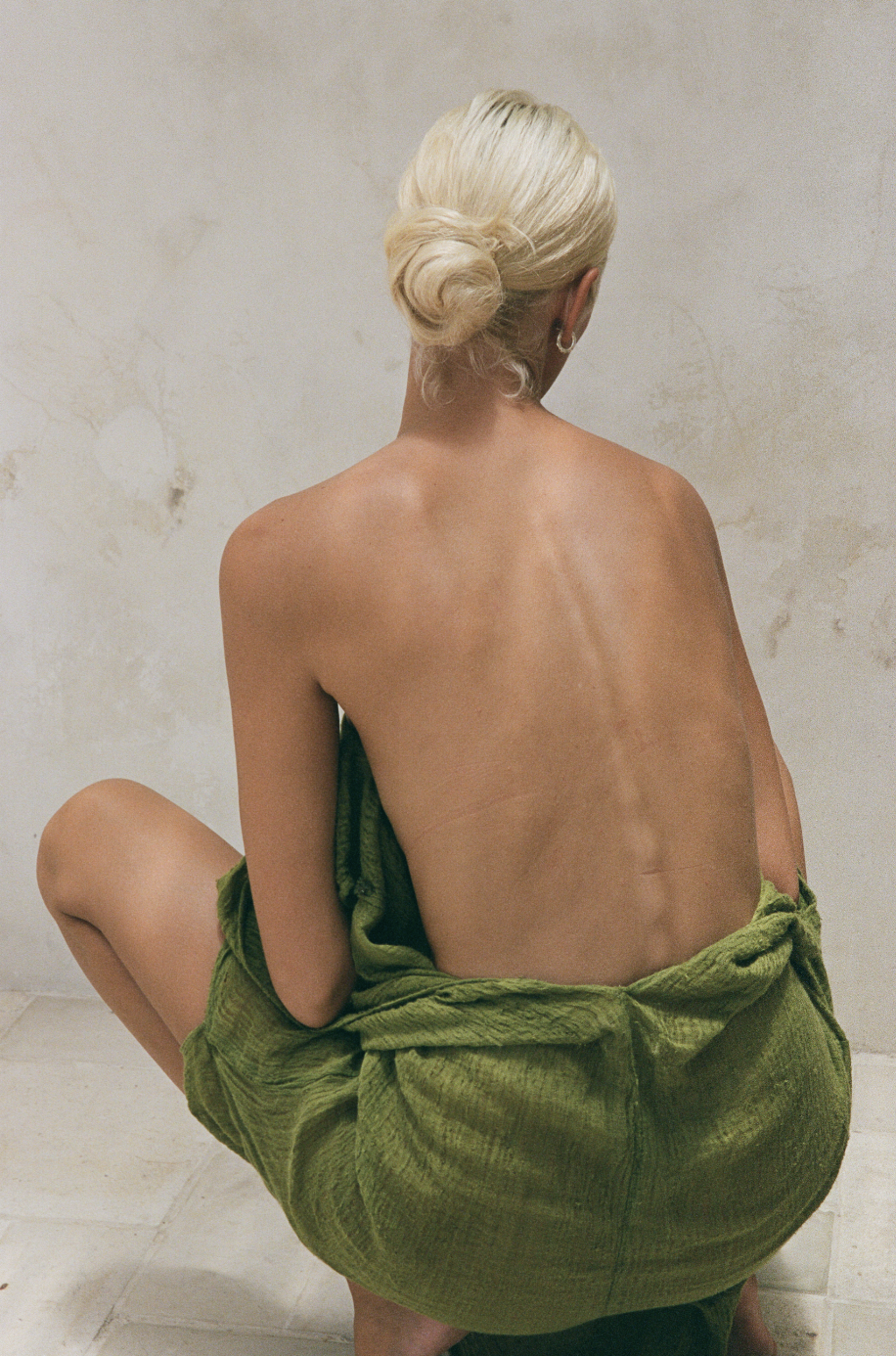 Person with blonde hair in a bun, seated and facing away, with a bare back and wrapped in a green textured fabric, against a neutral background.