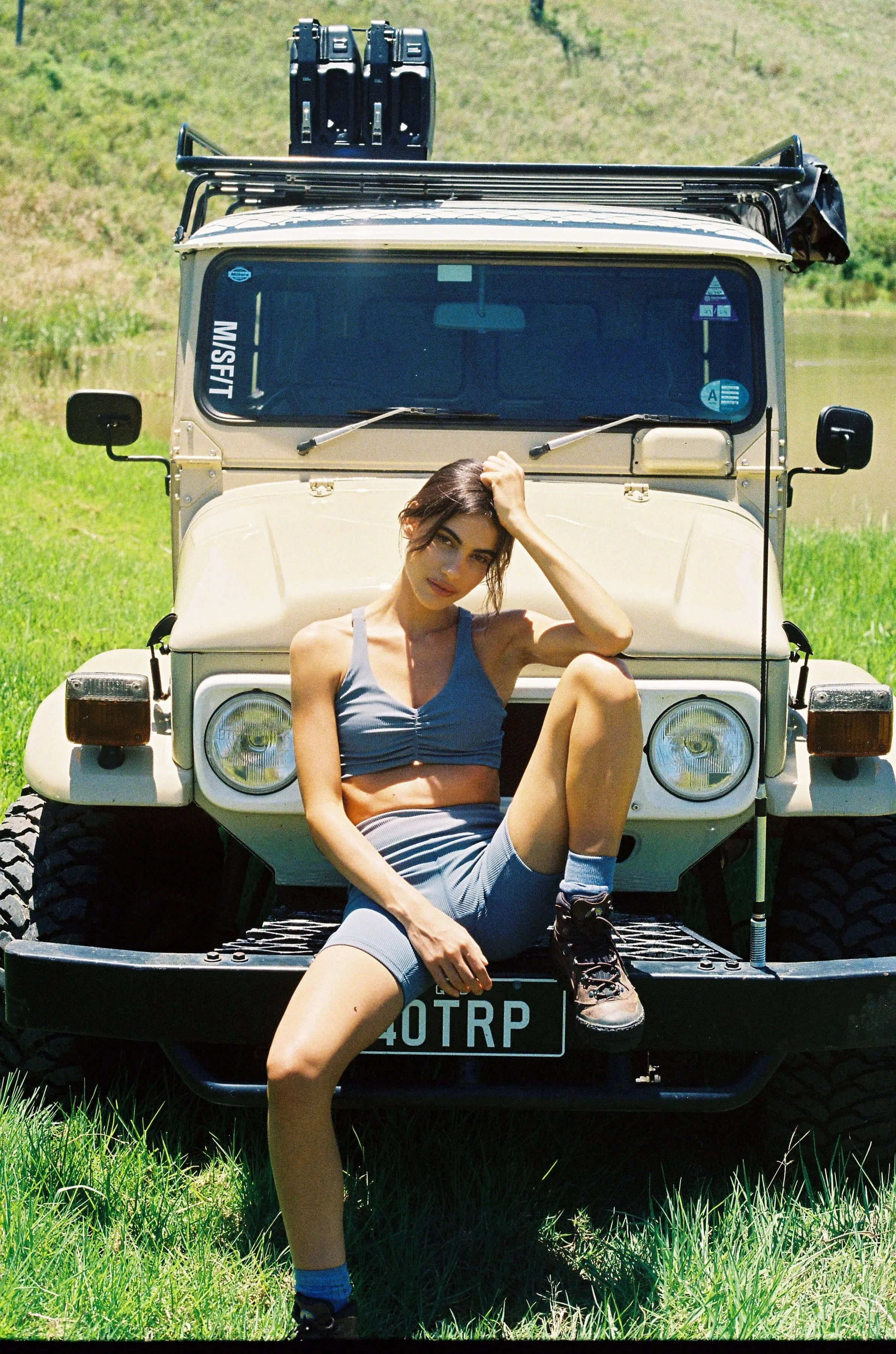 Person in athletic wear sitting on the front bumper of a beige vintage off-road vehicle parked on grass, with luggage on the roof and a pond in the background.