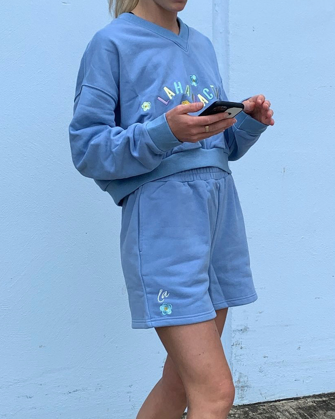 Person wearing a blue sweatshirt and shorts, holding a smartphone, standing against a light blue wall.