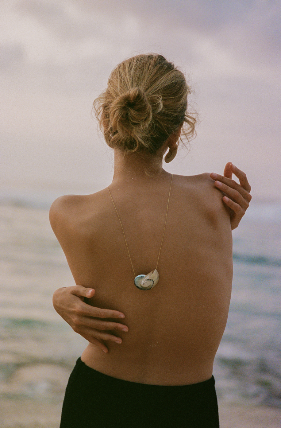Woman with blonde hair in a bun, facing the ocean, wearing a necklace with a pendant on her back.