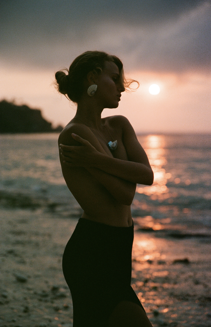 Silhouette of a woman on a beach at sunset, wearing a dark skirt and large earrings, with her arms crossed over her chest. Photoshoot for Olami Jewellery in Bali.