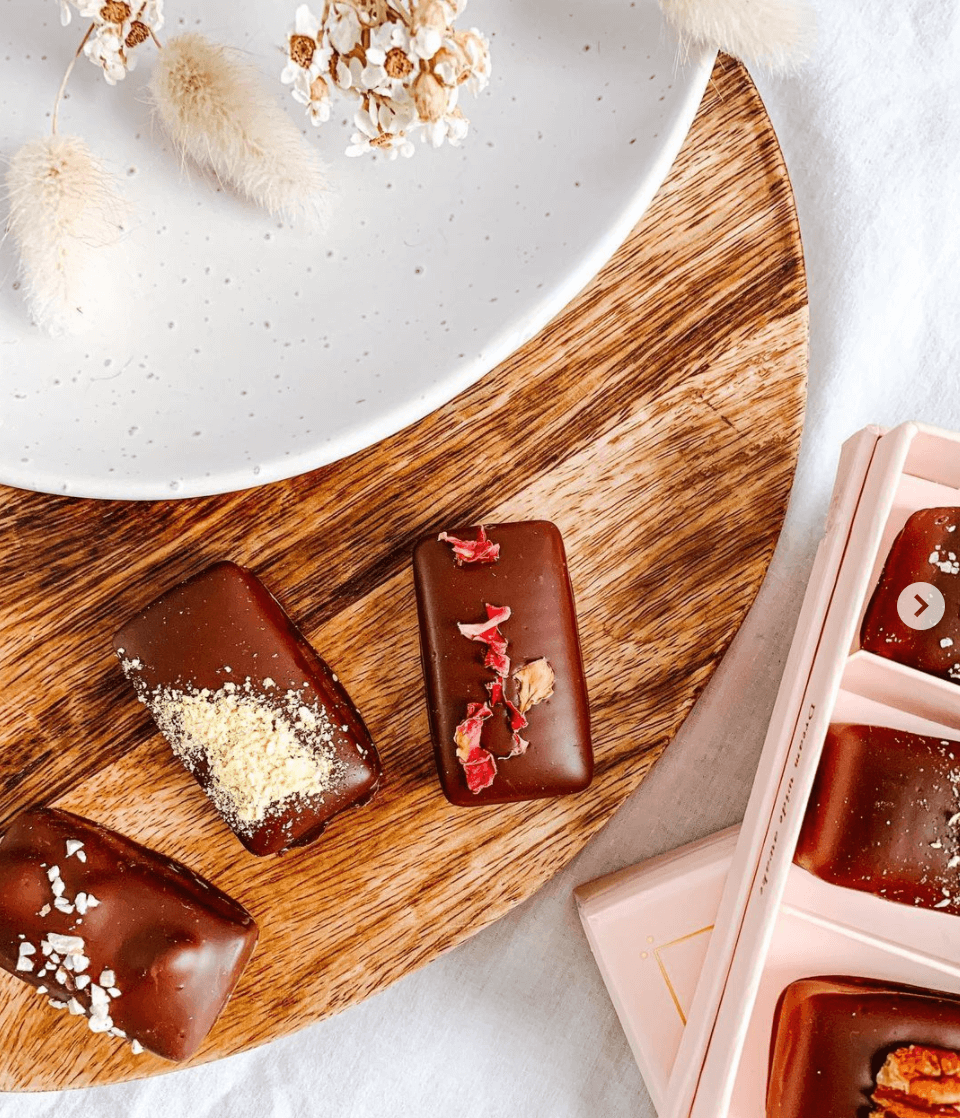 Assorted chocolate bars with toppings on a wooden board, white plate with dried flowers, and a pink chocolate box on the side.