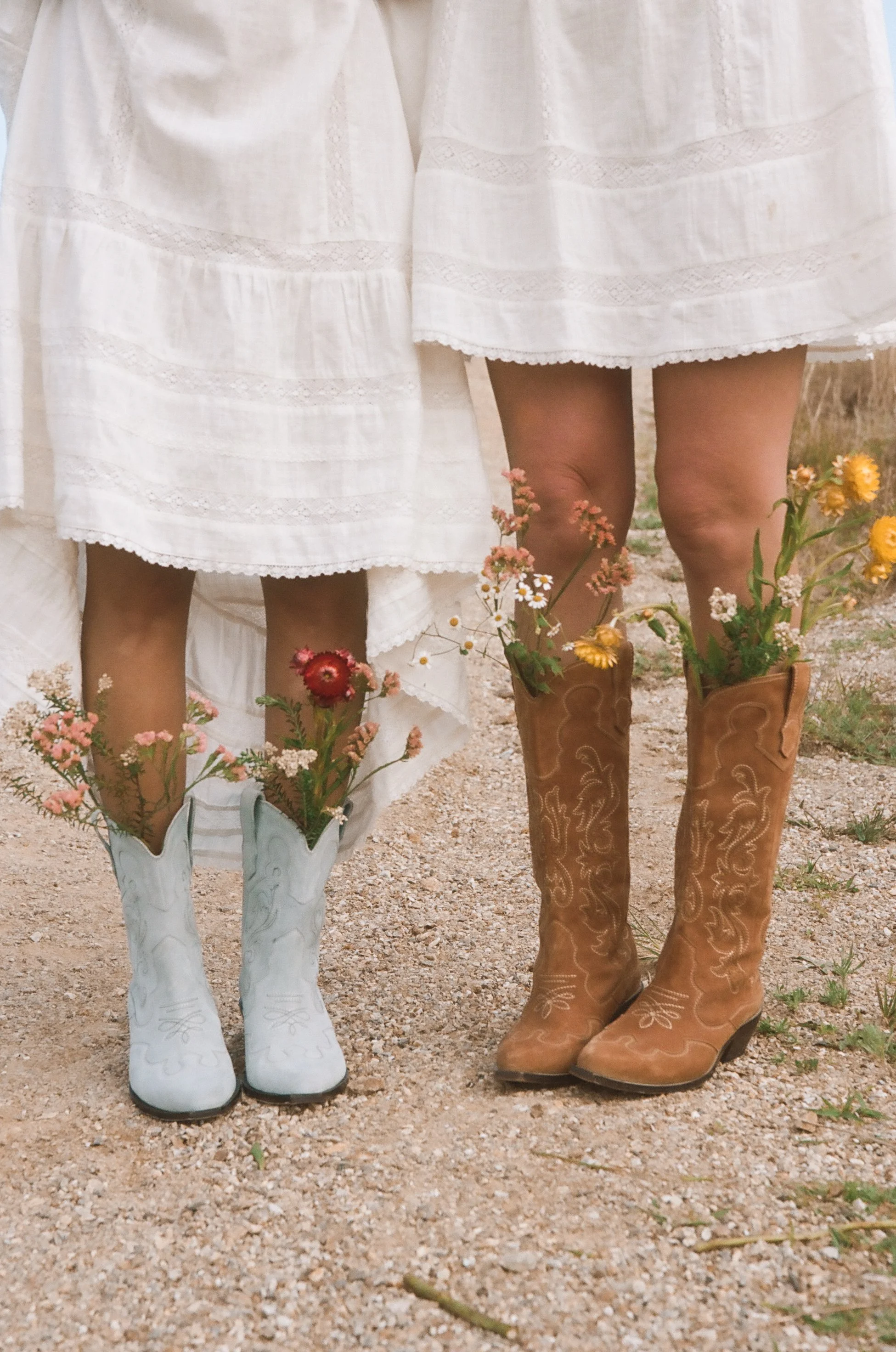 Two women wearing white dresses and cowboy boots filled with wildflowers standing on a gravel path.