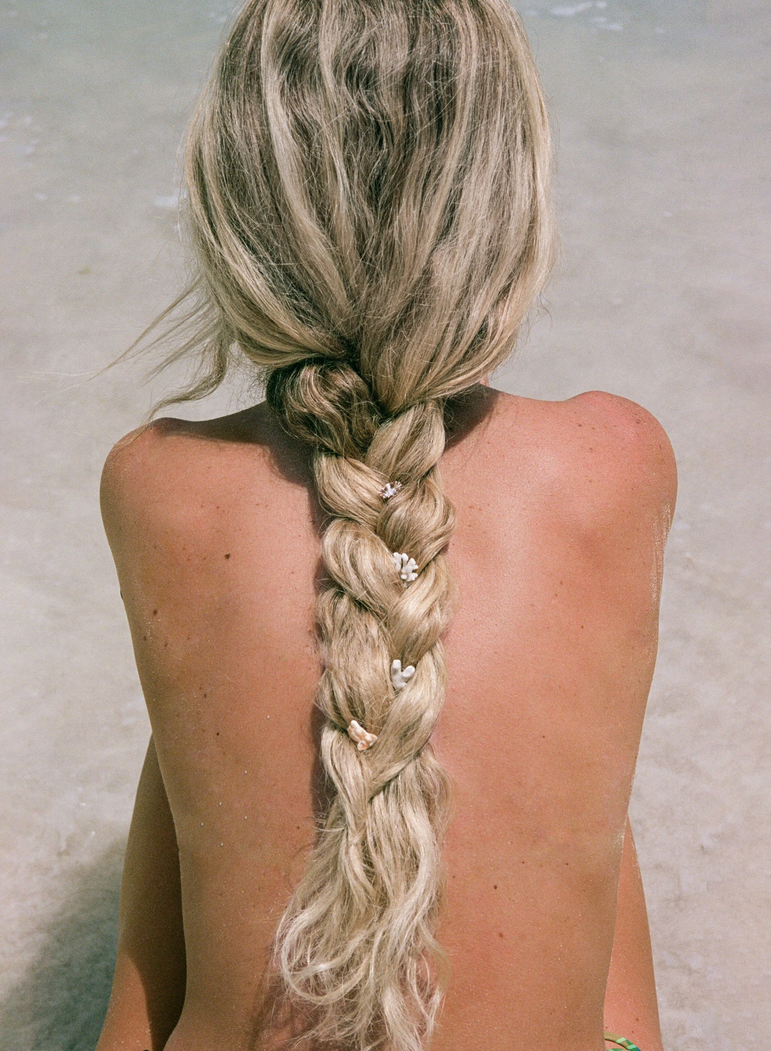 Person with long blonde braid sitting on a sandy beach.