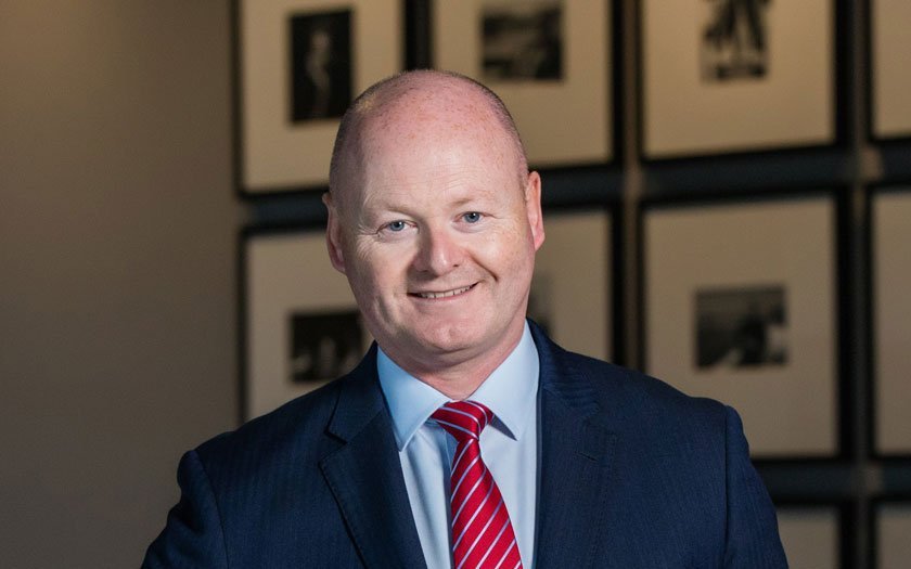 A middle-aged man with a bald head wearing a dark suit, light blue shirt, and red and white striped tie, smiling in front of a wall with framed black-and-white photographs.