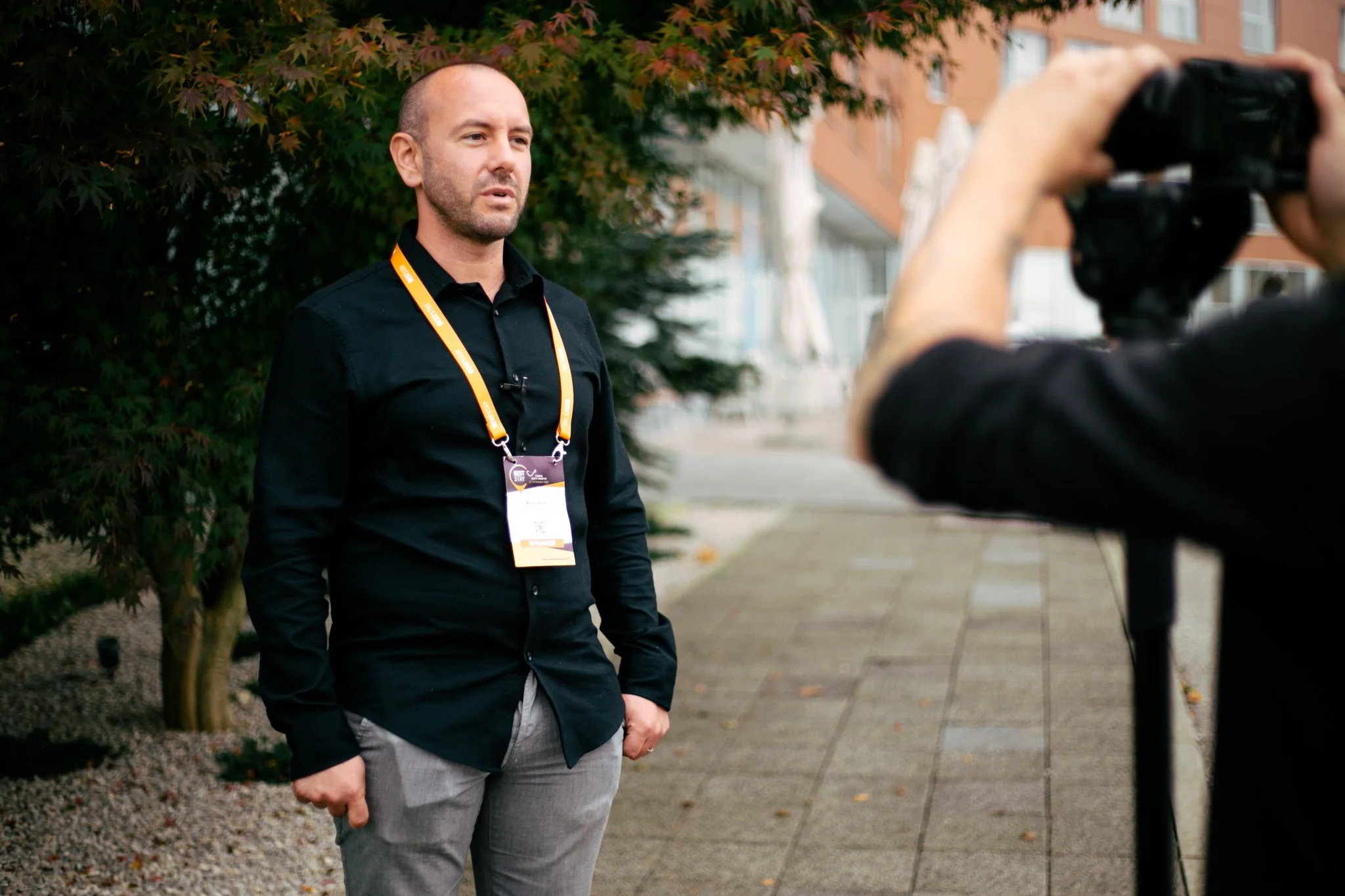 Man being interviewed outdoors, wearing a black shirt and grey pants, with an orange lanyard and a badge, standing on a sidewalk with trees and buildings in the background.