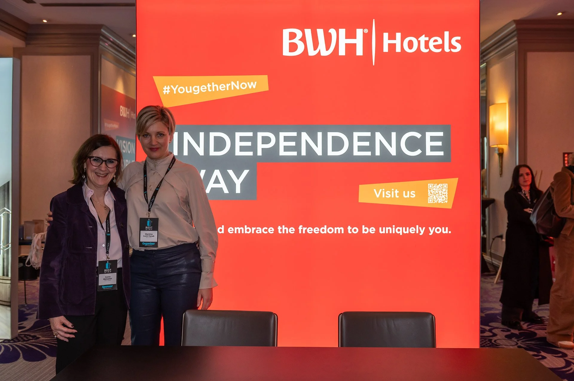 Two women standing together in front of a large red banner at a conference. The banner displays 'BWH Hotels' and 'Independence Day' with hashtags and a QR code.