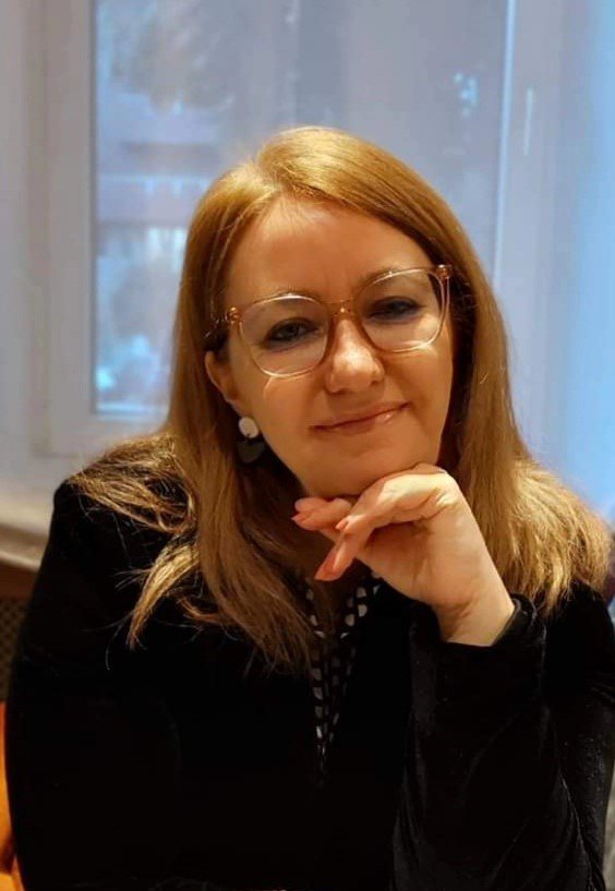 A woman with shoulder-length blond hair and glasses, smiling with her chin resting on her hand, sitting in front of a window.