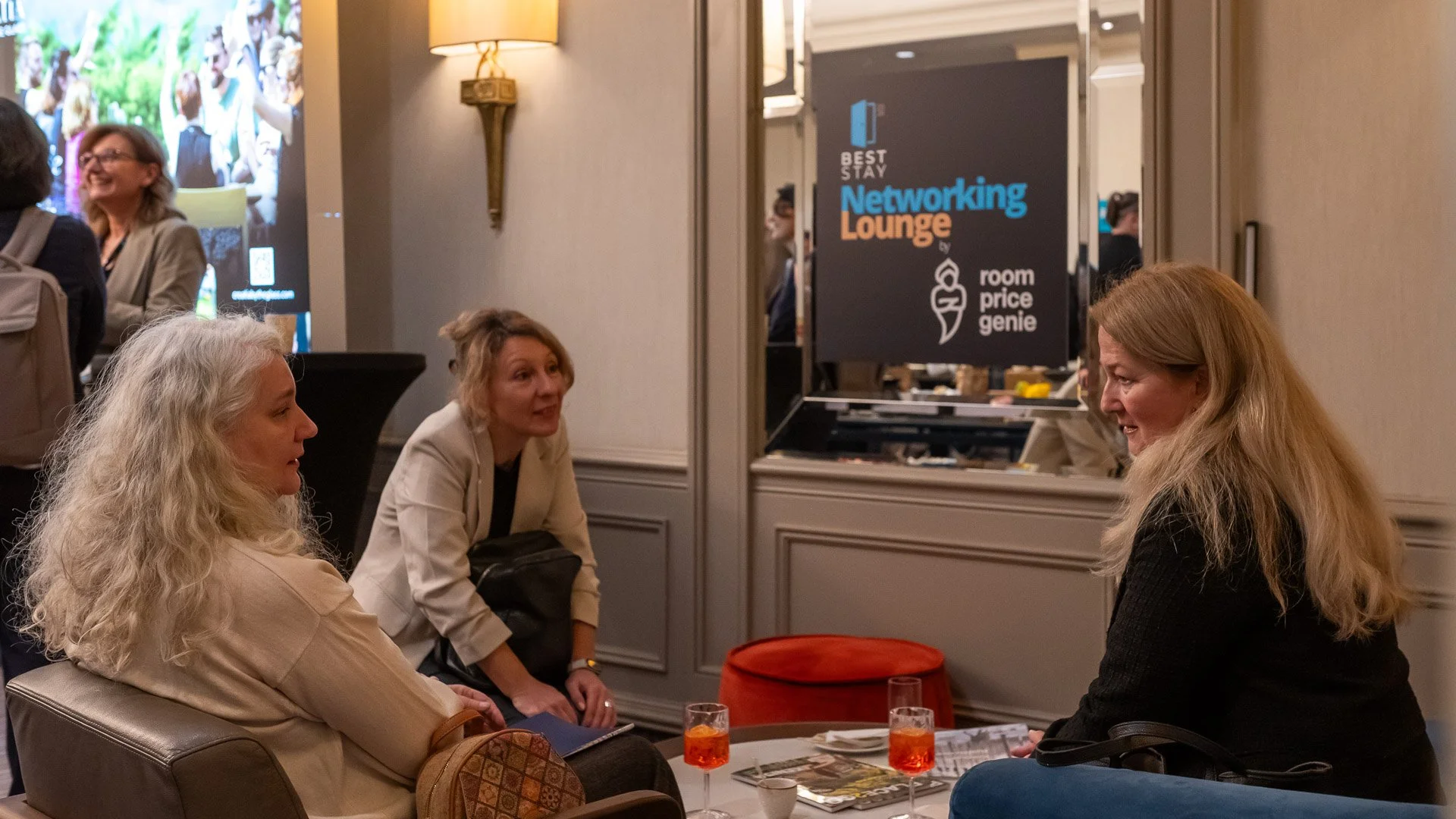 Three women sitting and talking at a networking lounge during a conference or event, with two glasses of orange-colored drinks on the table in front of them.