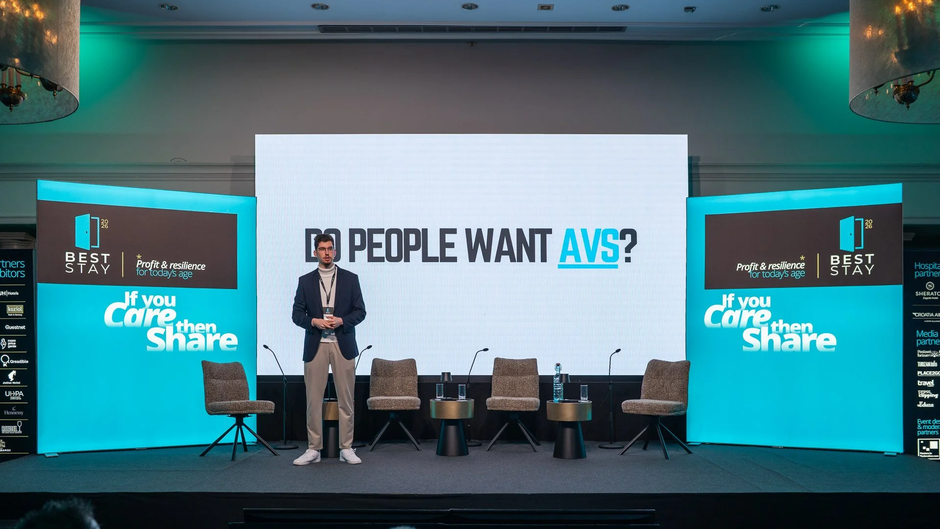 A man standing on stage in front of a large screen with text asking 'Do people want AVS?' at a conference or event. There are four chairs and small tables on stage, with water bottles on the tables. The background has blue and black branding with add