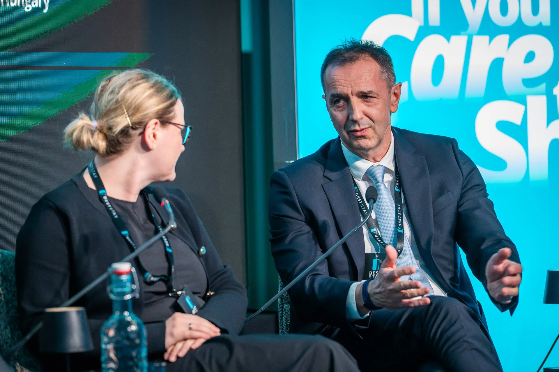 A man and a woman are seated on stage at a conference or panel discussion, engaged in conversation. The man wears a dark suit and light blue tie, gesturing with his hands while speaking. The woman wears glasses, a dark blazer, and a black top, listen