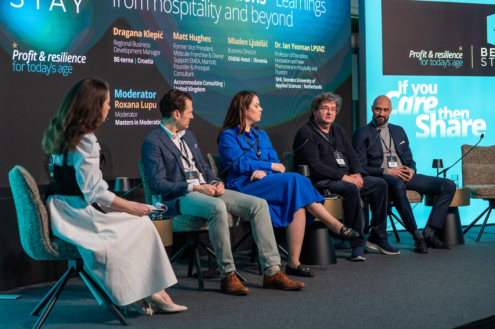 A panel discussion with six people seated on stage at a conference, against a backdrop displaying text and names of speakers.