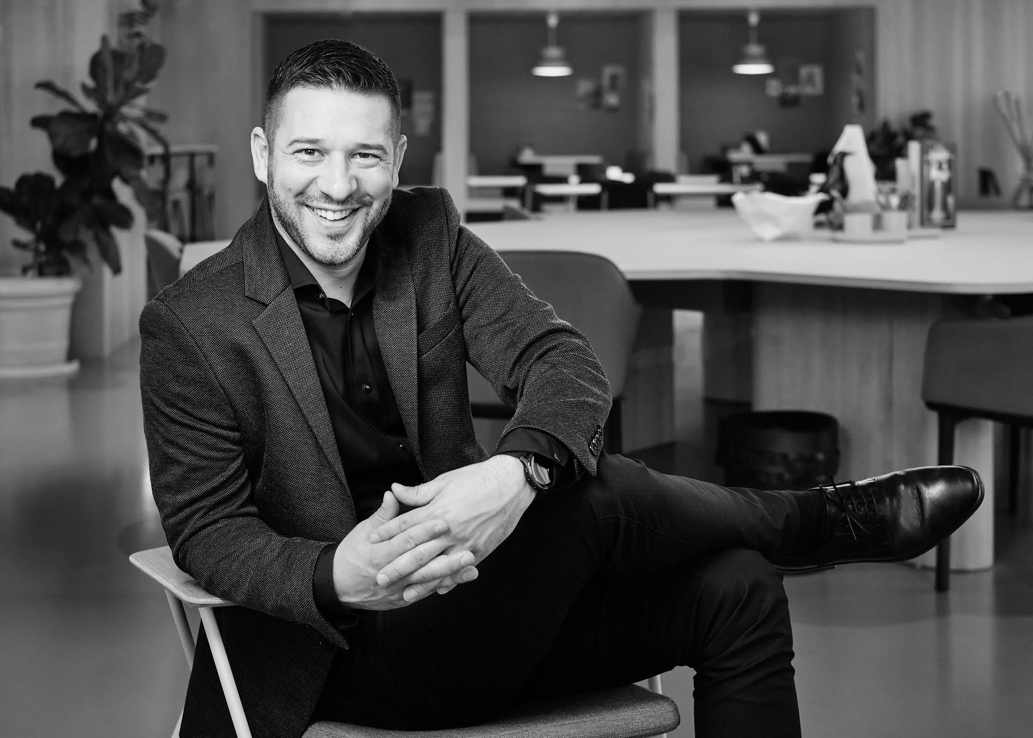 A smiling man in business casual attire sitting on a chair in an office or coworking space, with a large table and plants in the background.