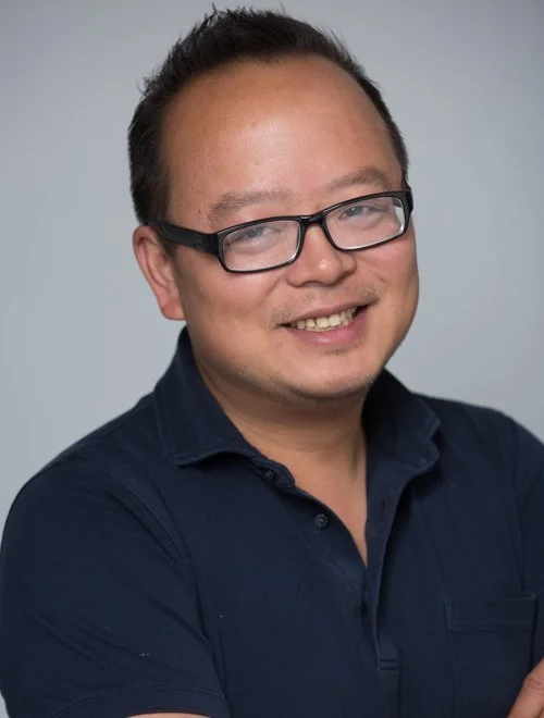 Headshot of a man wearing glasses and a dark blue collared shirt, smiling with arms crossed against a plain light gray background.
