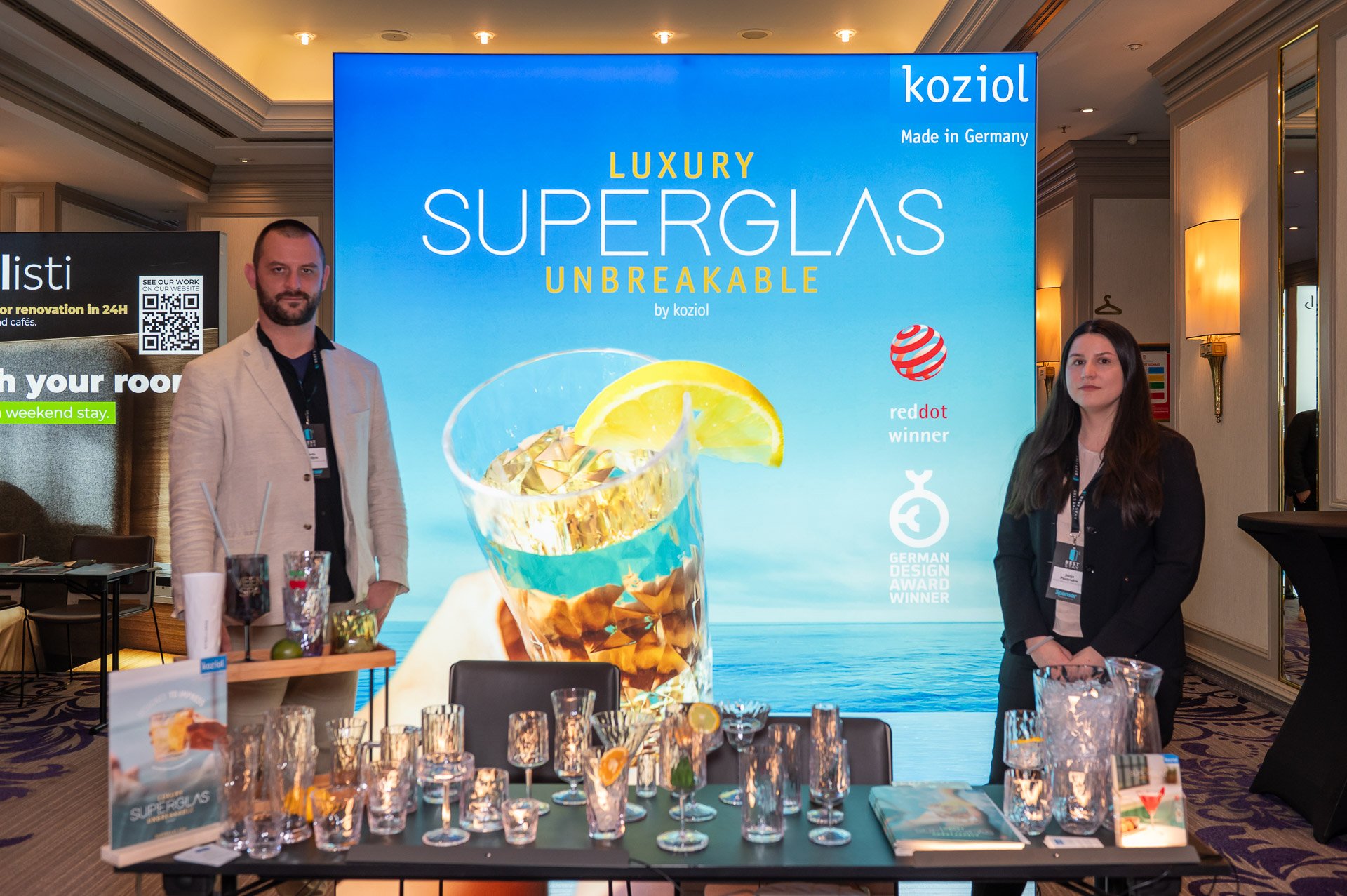 Two people standing in front of a large blue advertising banner for Kozioł superglas, with a table of glasses and promotional materials in the foreground.