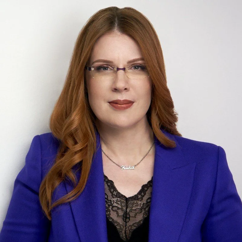 A woman with long red hair, wearing glasses and a blue blazer, standing against a plain white background.