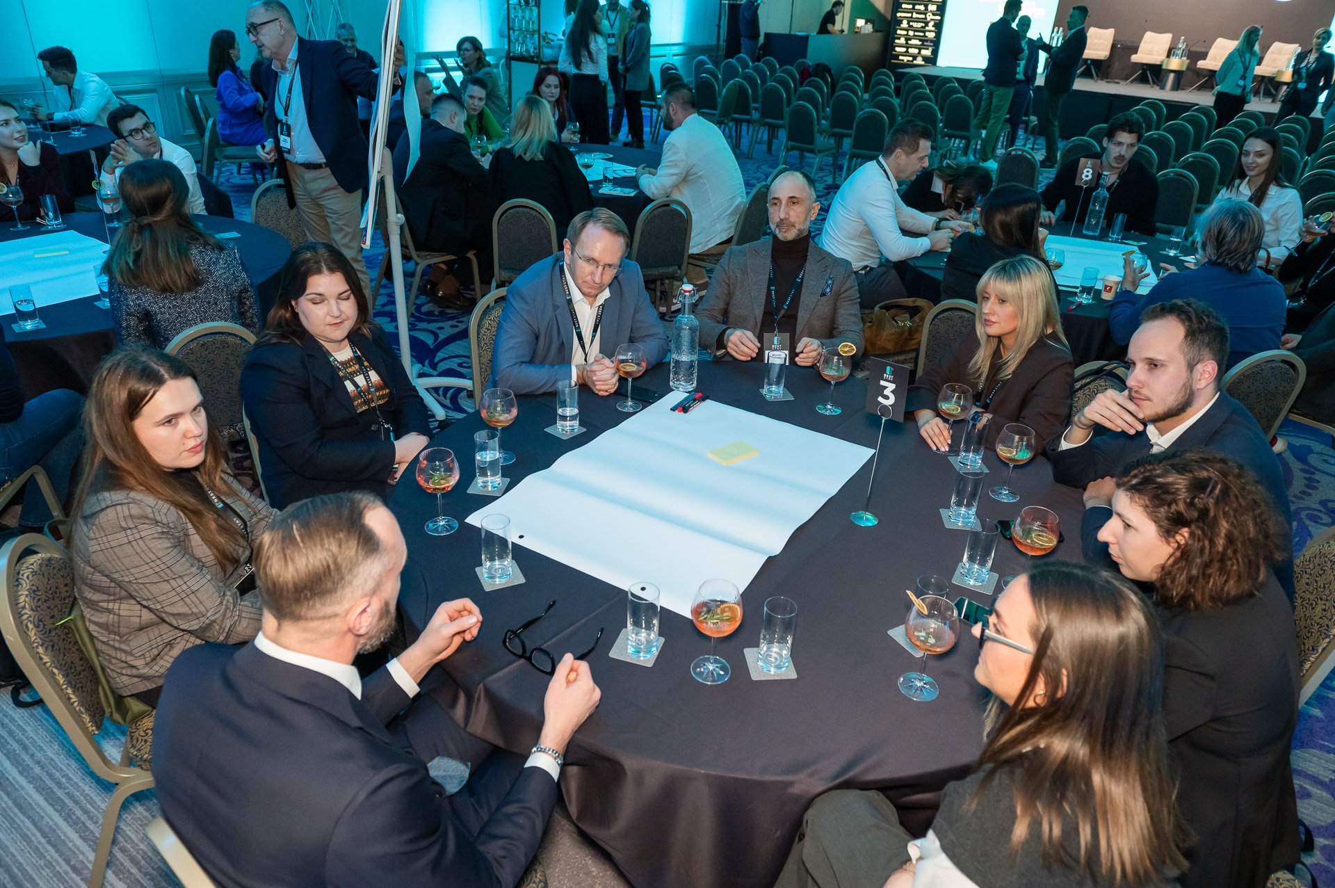 Group of people in business attire seated around a circular table at a conference or networking event, engaged in conversation, with glasses of wine and water on the table.