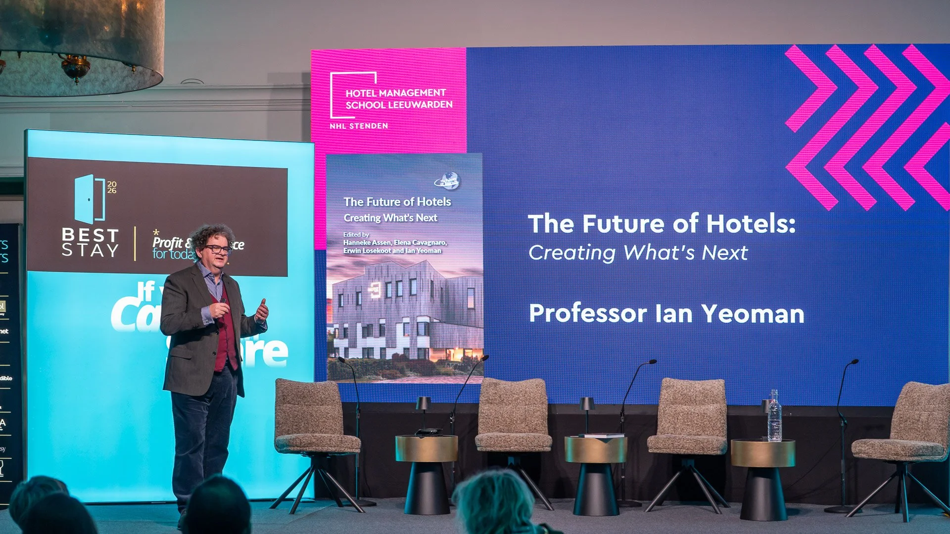 A man wearing a suit and glasses stands on a stage, speaking at a conference. Behind him are multiple large screens displaying text and images related to hotels and hotel management. There are empty chairs on the stage, with one holding a water bottl
