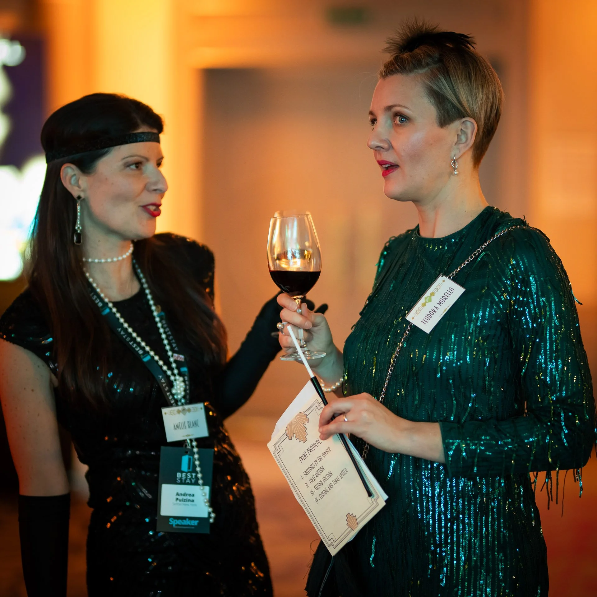 Two women dressed in vintage, glamorous attire talking at a formal event; one holds a glass of red wine, and the other holds a document and a glowing wand.