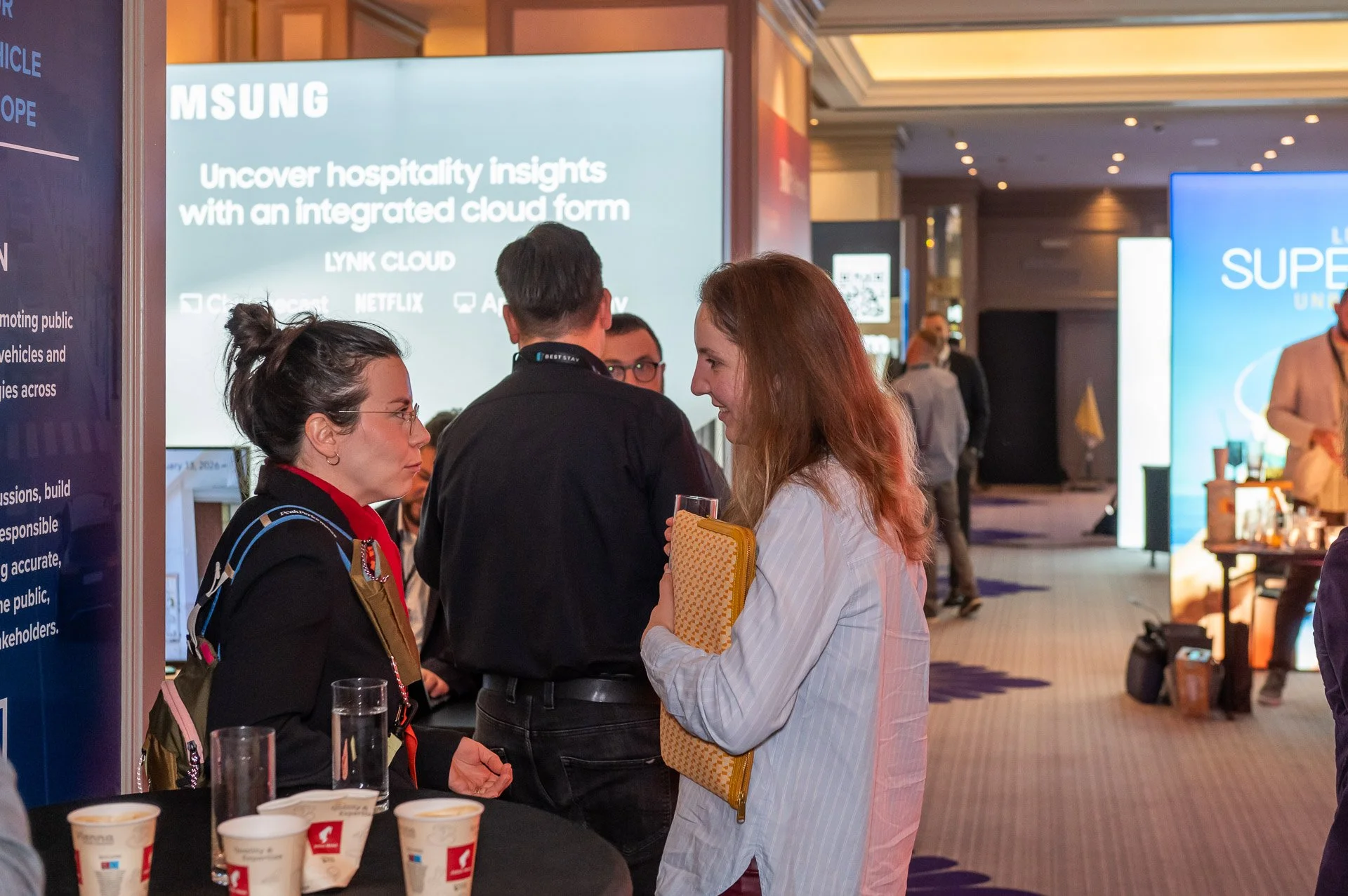 A woman with glasses and a backpack converses with another woman holding a yellow notebook and a glass of water at a conference or trade show, with large display screens and other attendees in the background.