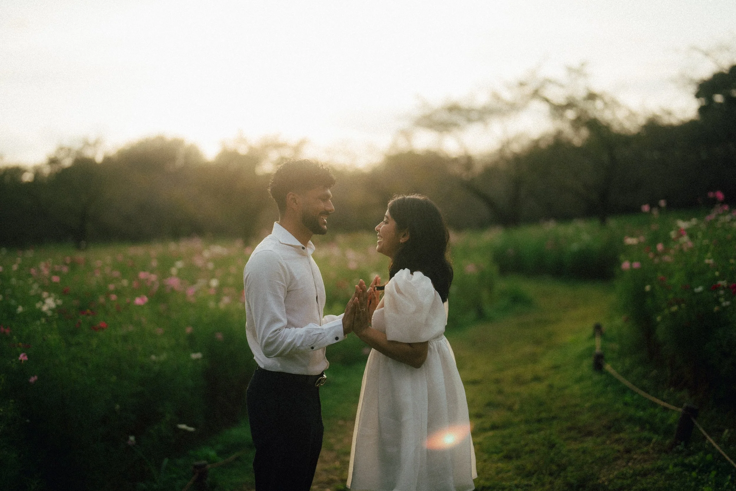 A couple standing close in a flower garden at sunset, smiling and holding hands, dressed casually, surrounded by blooming flowers and trees in the background.
