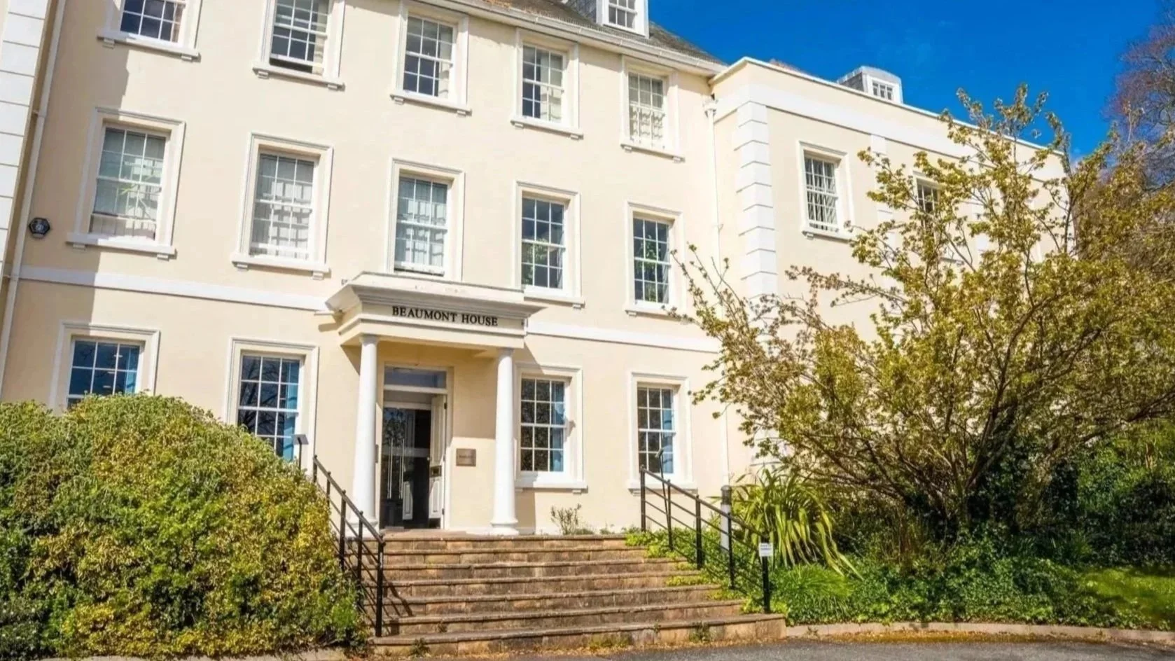 A multi-story white building labeled 'Beaumont House' with a front porch supported by two white columns, stone steps leading up to the entrance, surrounded by green bushes and trees under a clear blue sky.