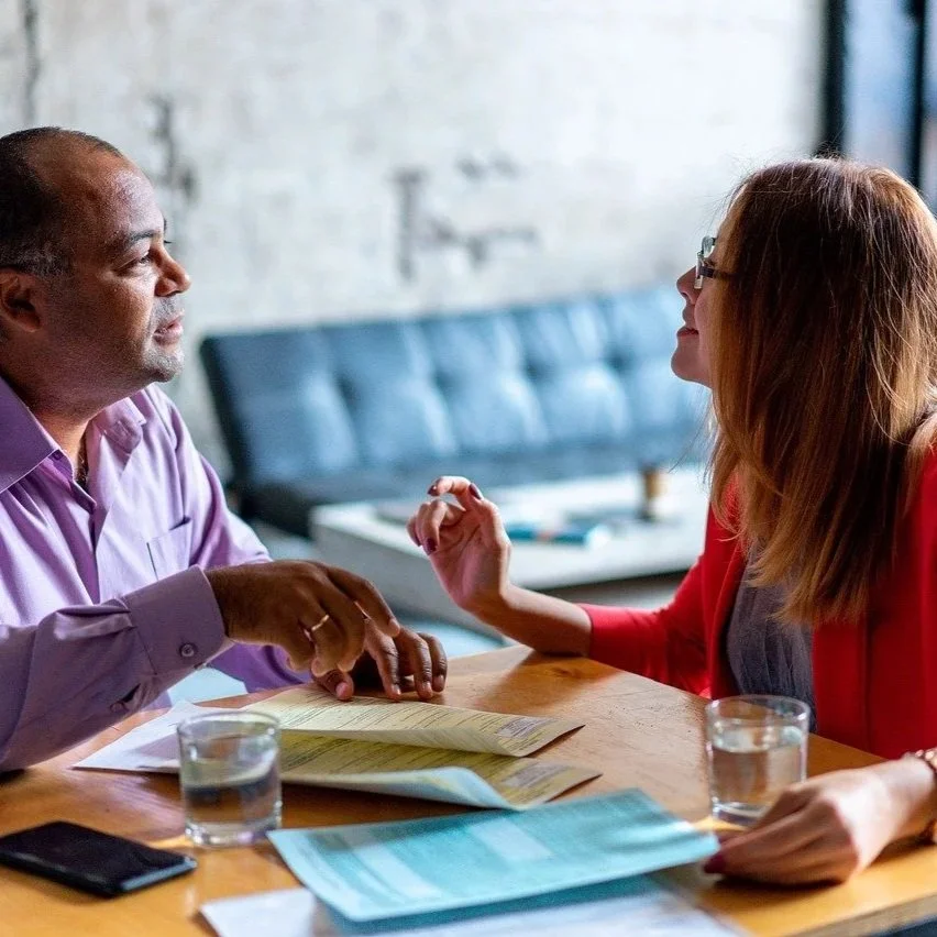Two people are talking at a table in a café; there are papers and glasses of water on the table, with a couch and a wall in the background.