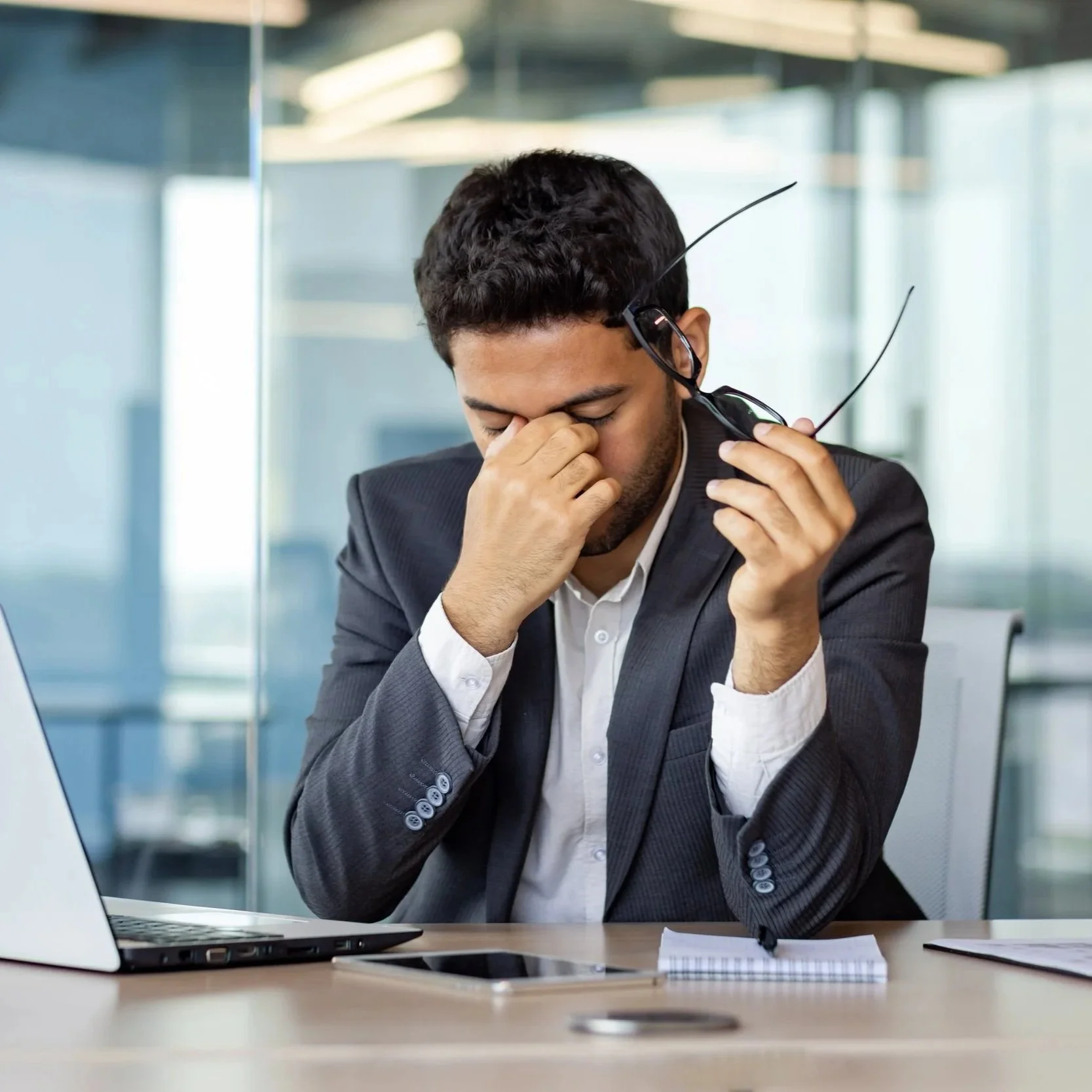 A businessman in a suit and glasses, sitting at a desk in an office, holding his nose and looking stressed or exhausted.