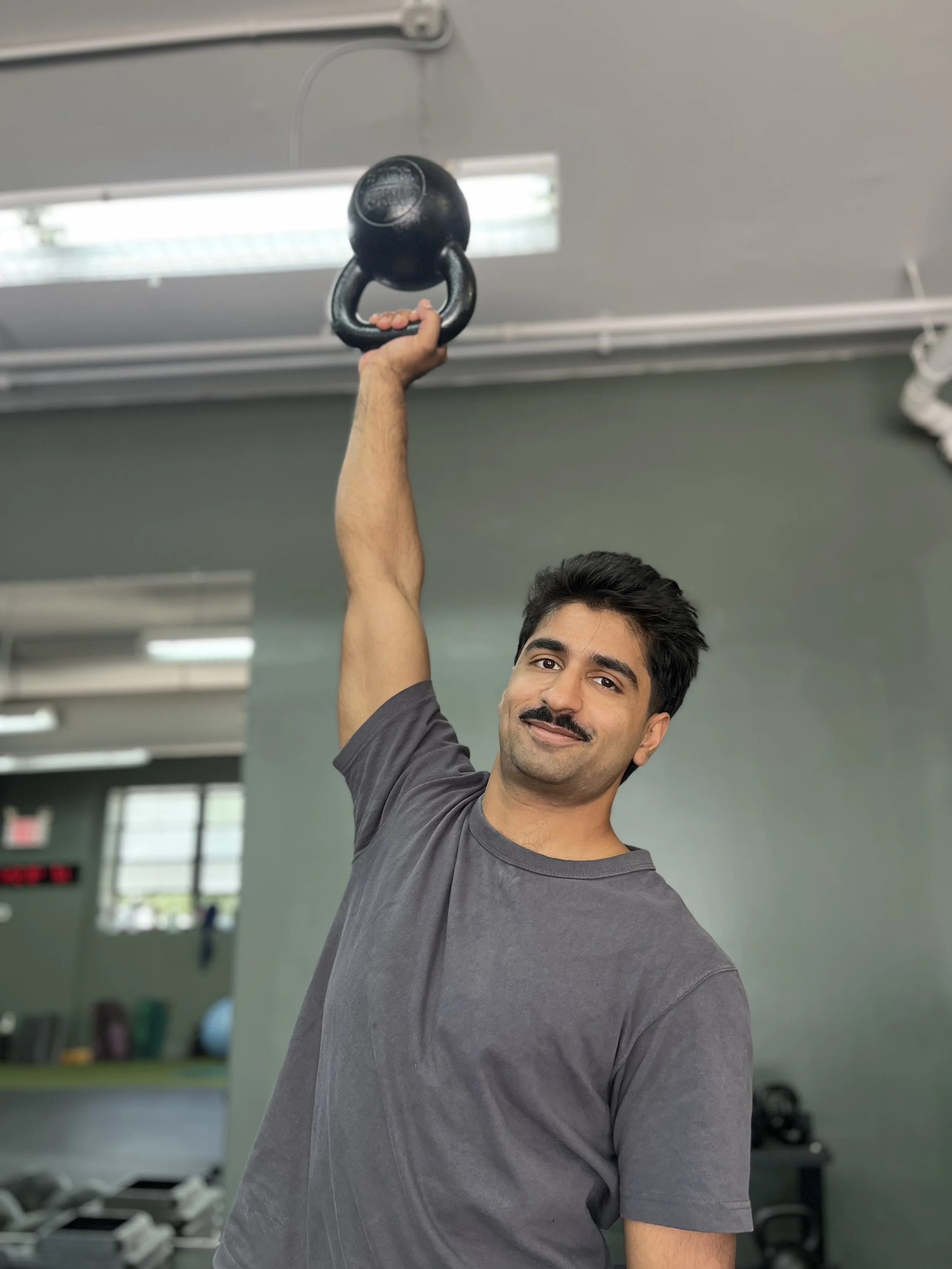 A man holds a black kettlebell in his right hand raised over his head in a gym setting.