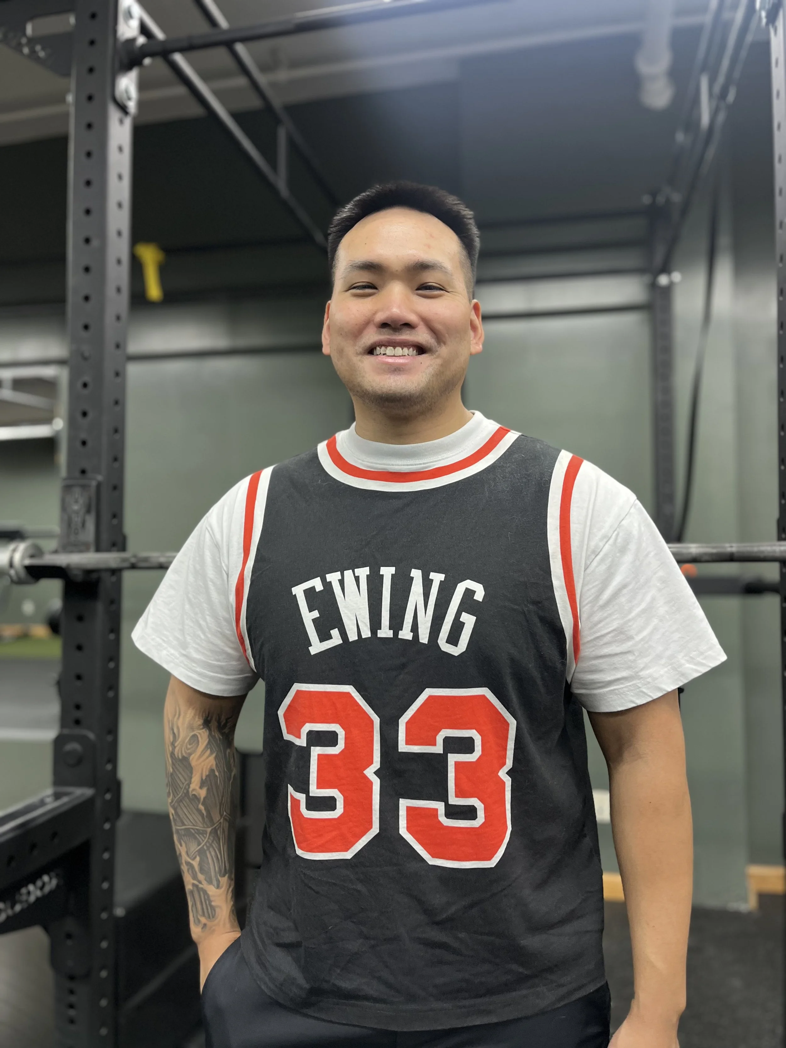 A man smiling at the camera inside a gym, standing near gym equipment, wearing a black basketball jersey with red and white details and the number 23.