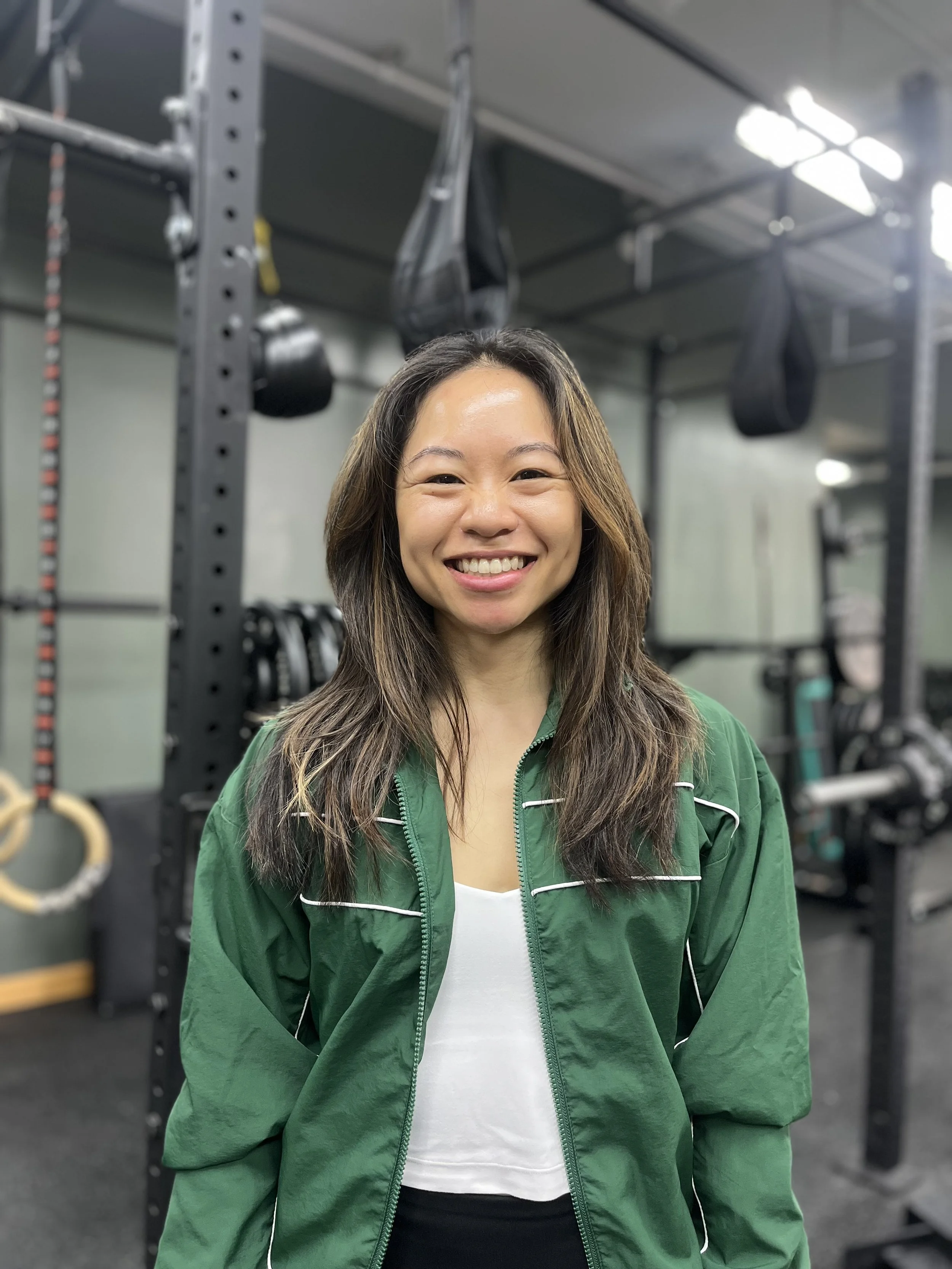 A smiling woman with long brown hair wearing a green jacket and white top standing in a gym with workout equipment in the background.
