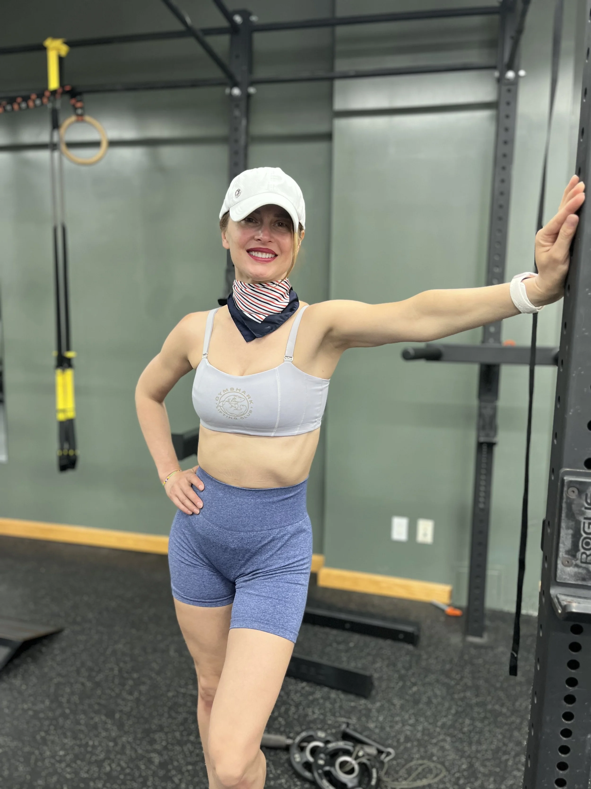 A woman with a white cap and sportswear posing in a gym, smiling and standing with one hand on her hip and the other resting on a gym rack.
