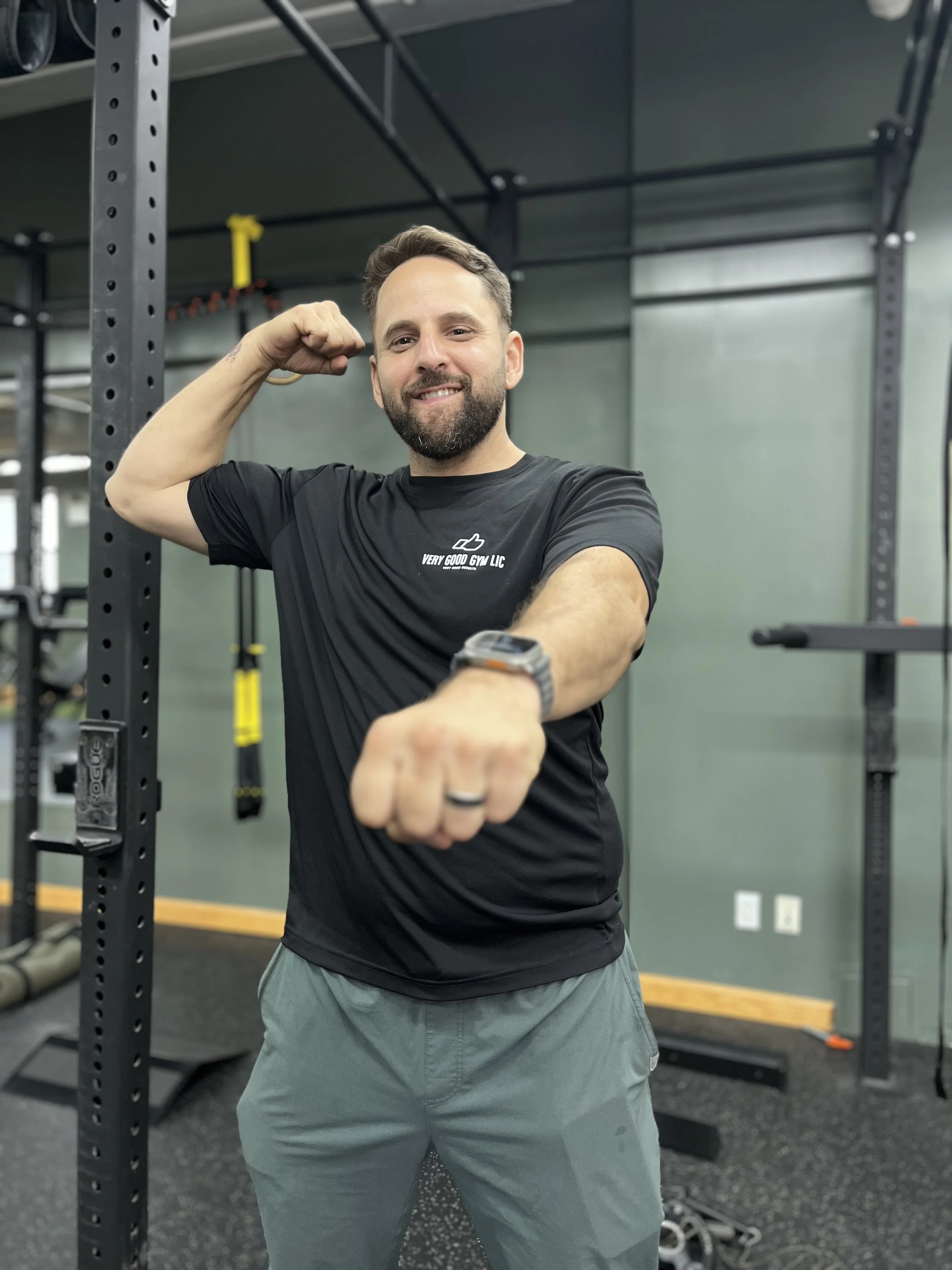A man flexing his arm at the gym, wearing a black T-shirt with a logo and text 'Very Good Gym LLC', and gray shorts, standing near gym equipment.