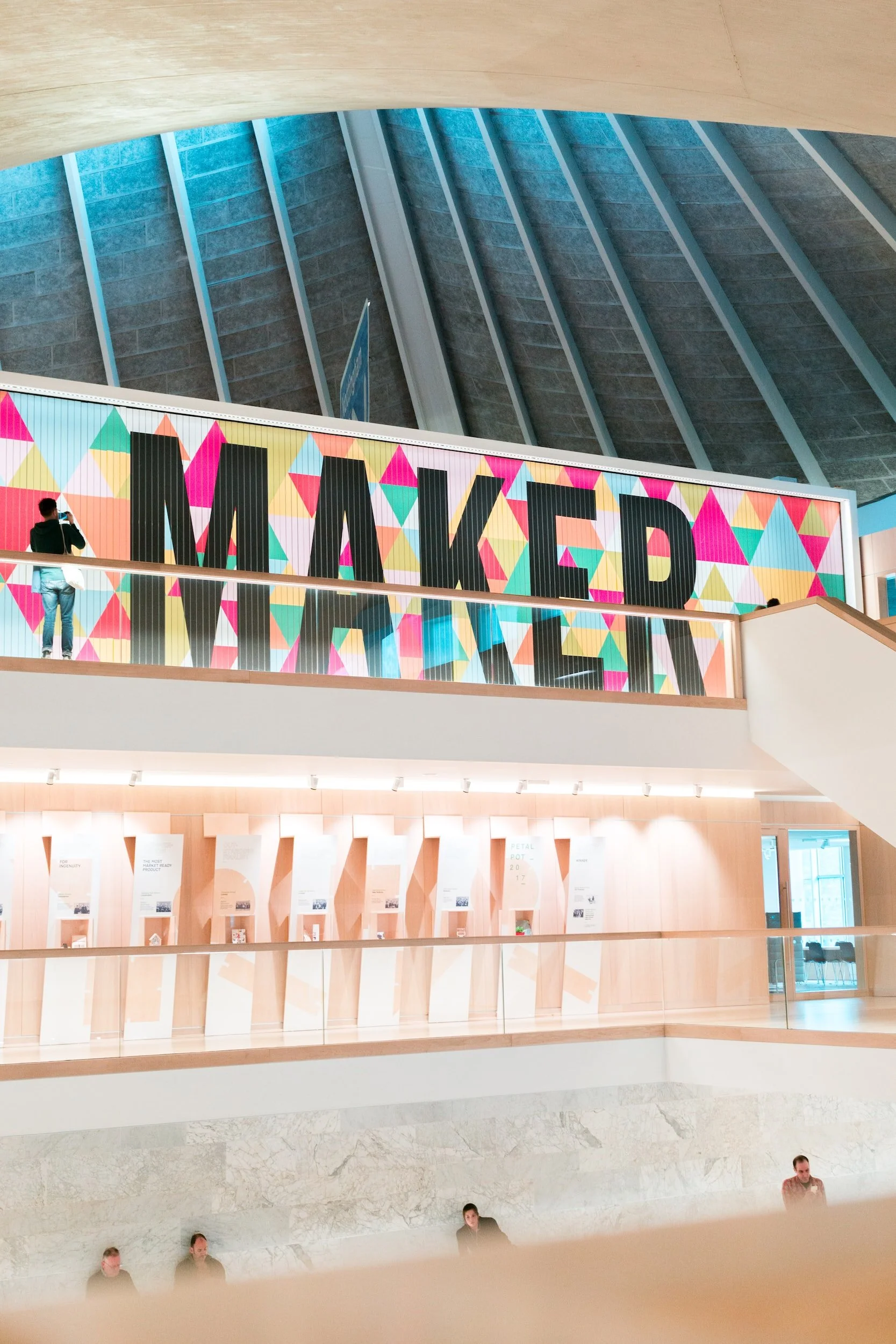 Interior of a modern building with a colorful mural that says 'MAKED' on an upper level, seen from below with several people inside.