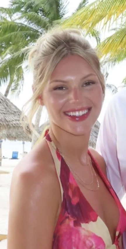 A blonde woman in a black polka-dot dress standing next to a hair sink in a salon.