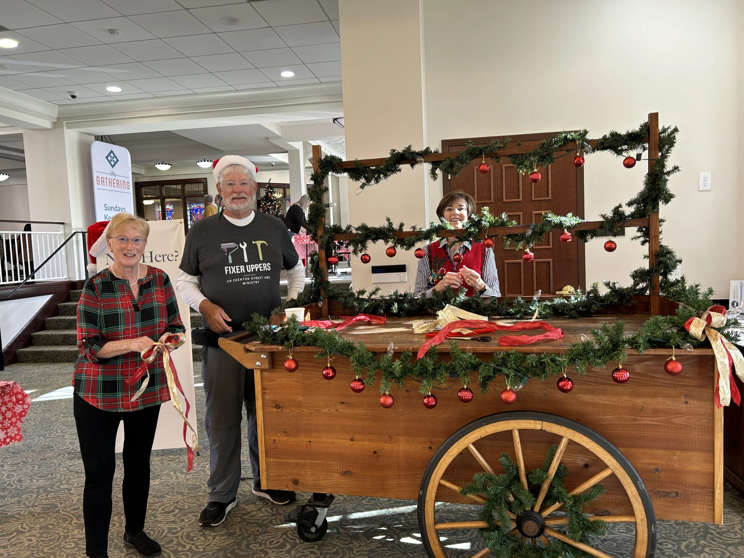 Three people decorating a wooden cart with Christmas garland and ornaments indoors. One man is wearing a "Fixer Uppers" shirt, and two women are holding ribbons. The setting appears festive with holiday decorations around.