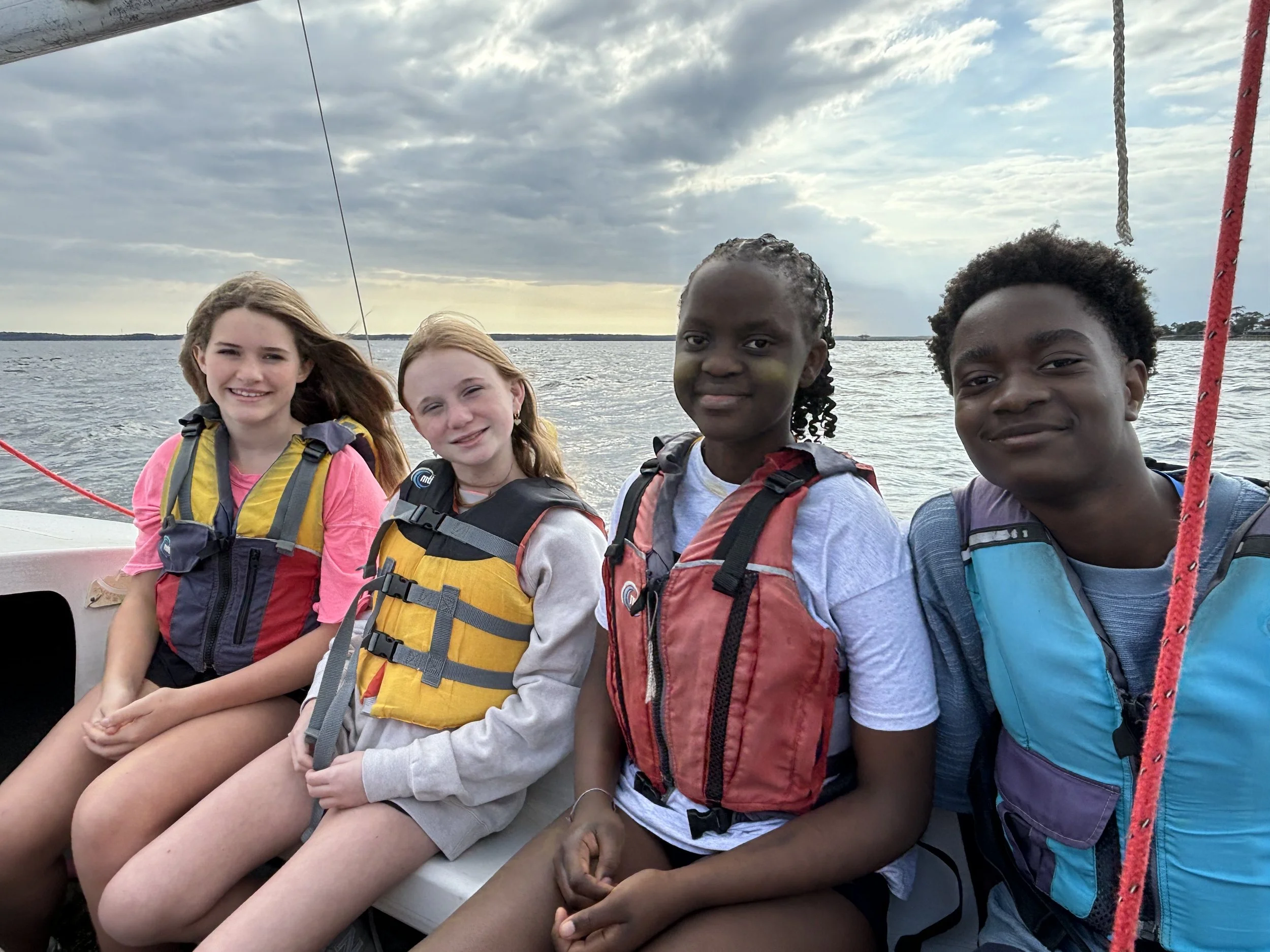 Four teenagers wearing life jackets sitting on a sailboat with water and cloudy sky in the background.