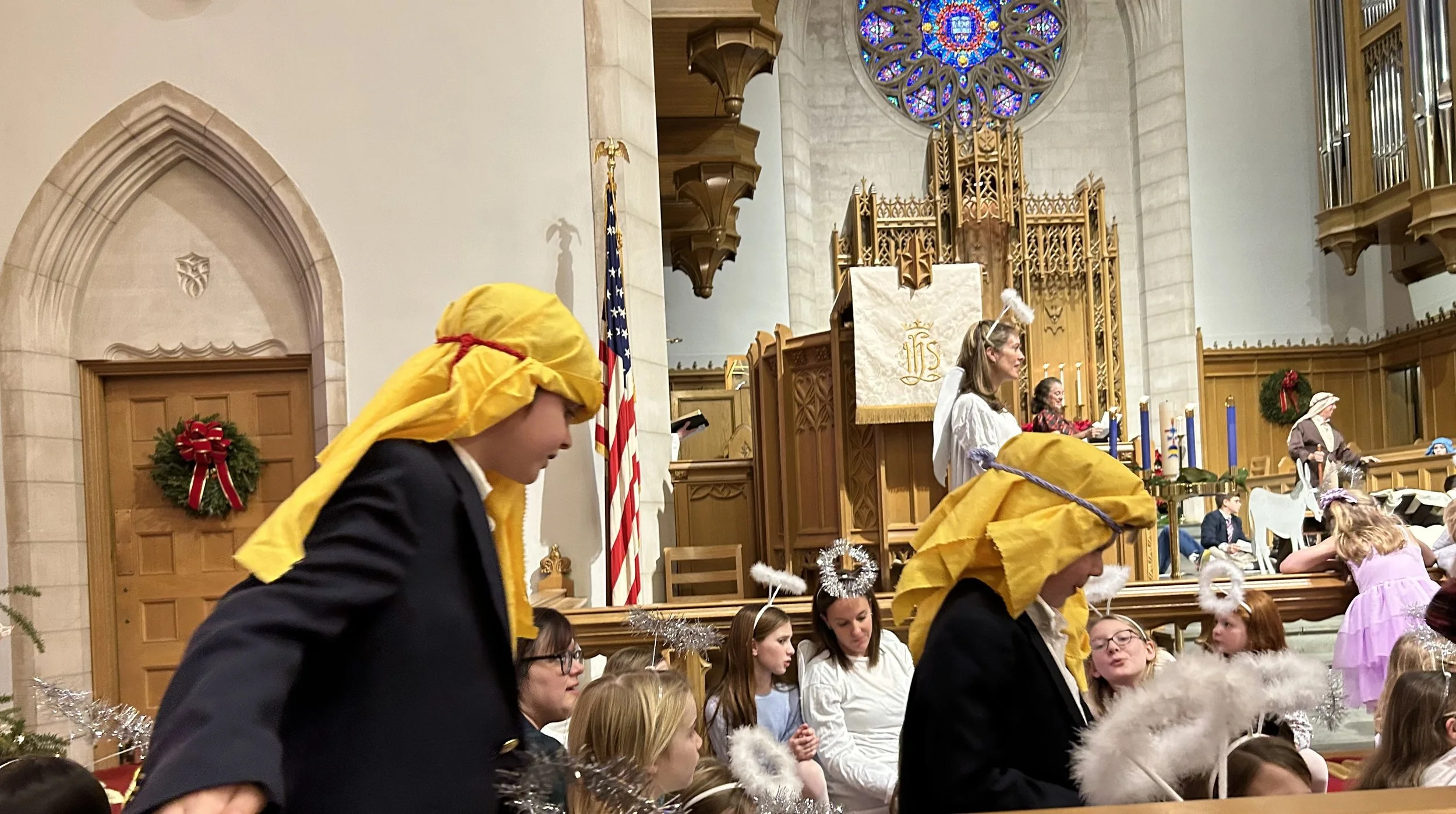 Children participating in a Christmas play at a church, wearing costumes such as yellow headscarves and angel wings. A decorated altar and stained glass window are visible in the background.