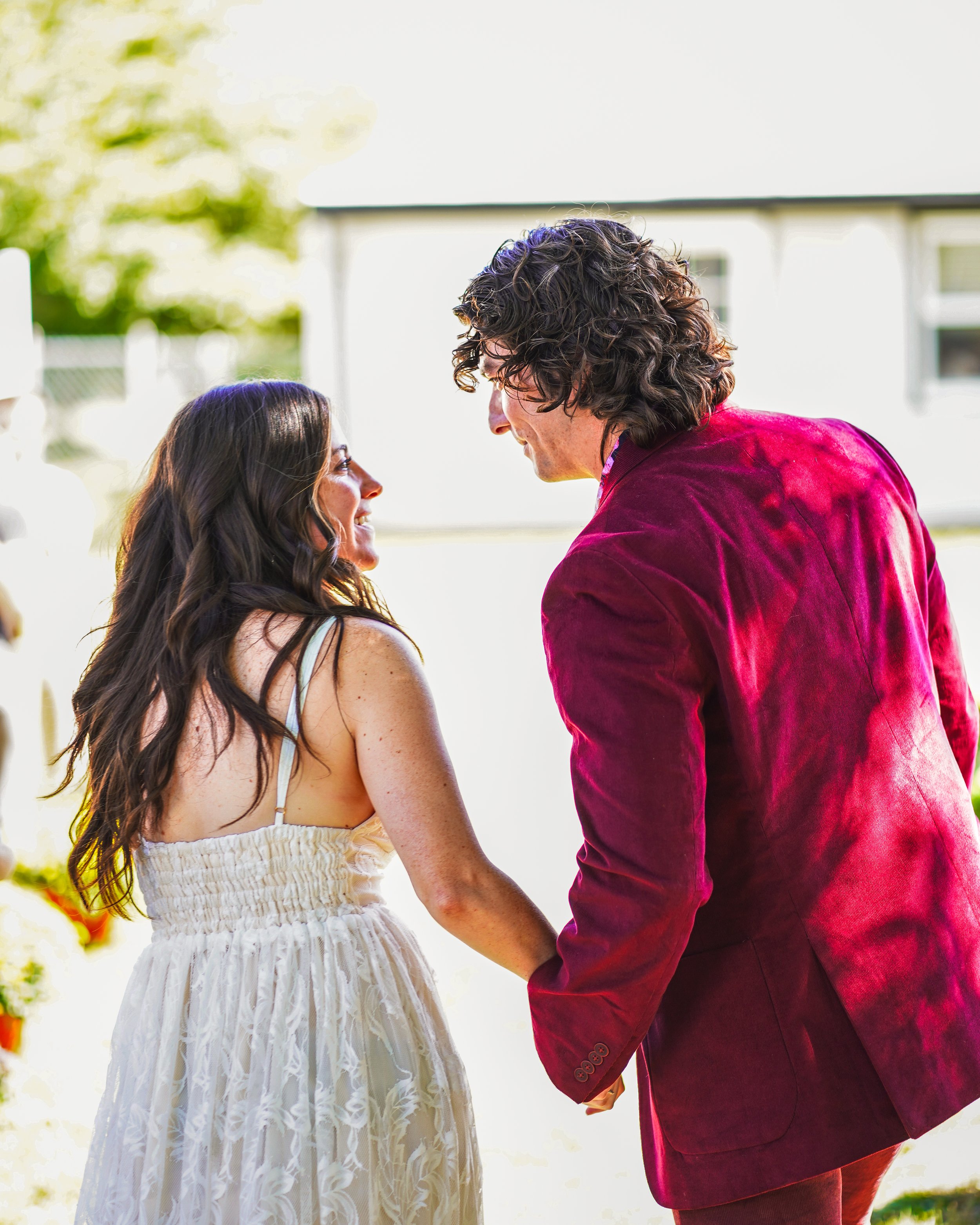 Couple holding hands outdoors, woman in white dress, man in red jacket.